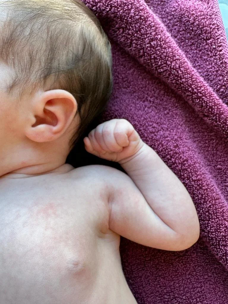 Close-up of a sleeping baby resting on a purple towel, showing part of the baby's head, arm, and hand clenched in a fist.