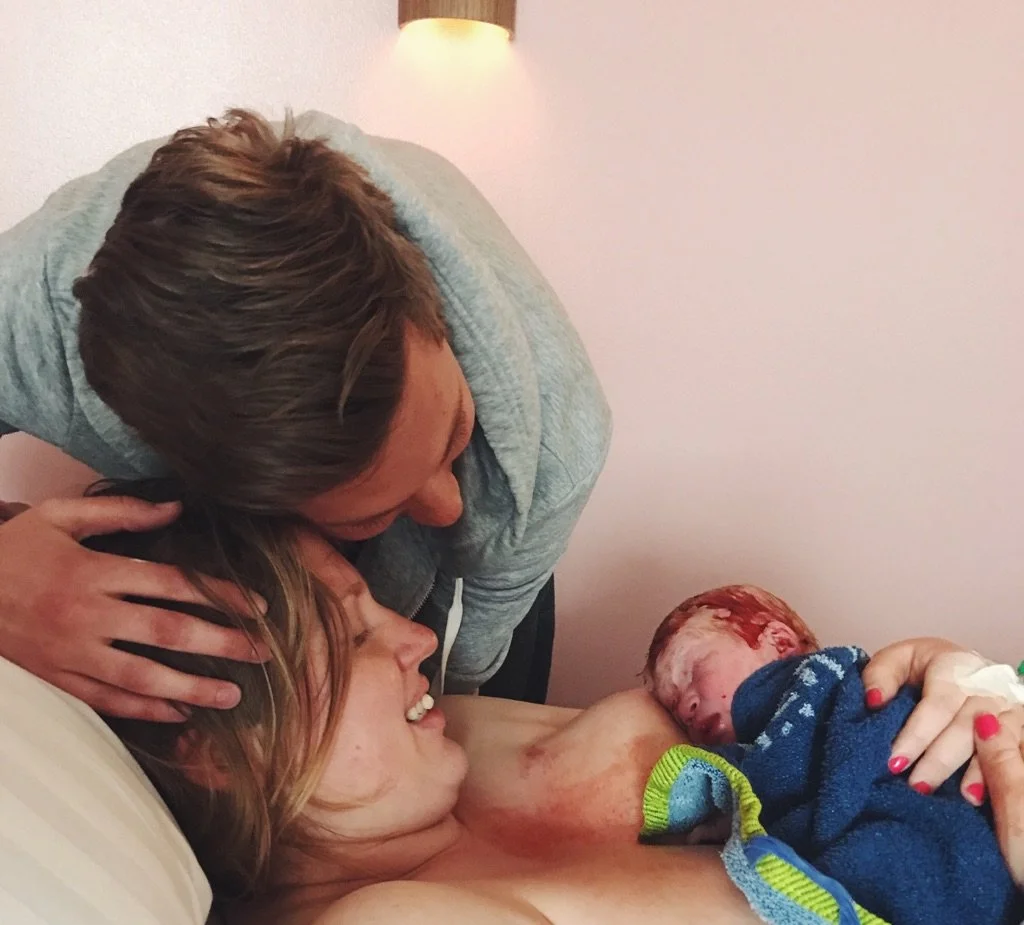 A woman and a man are with a newborn baby in a hospital room. The woman is lying in a hospital bed, holding the baby, and smiling. The man is leaning over, holding the woman's head, and kissing her forehead. The baby has a reddish appearance and is wrapped in a blue blanket.