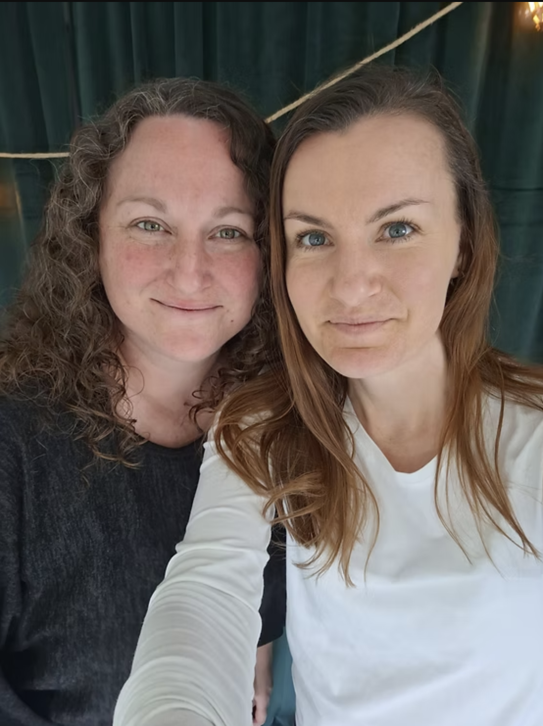 Two women taking a selfie together, smiling. The woman on the left has curly brown hair and is wearing a dark top. The woman on the right has straight auburn hair and is wearing a white top.
