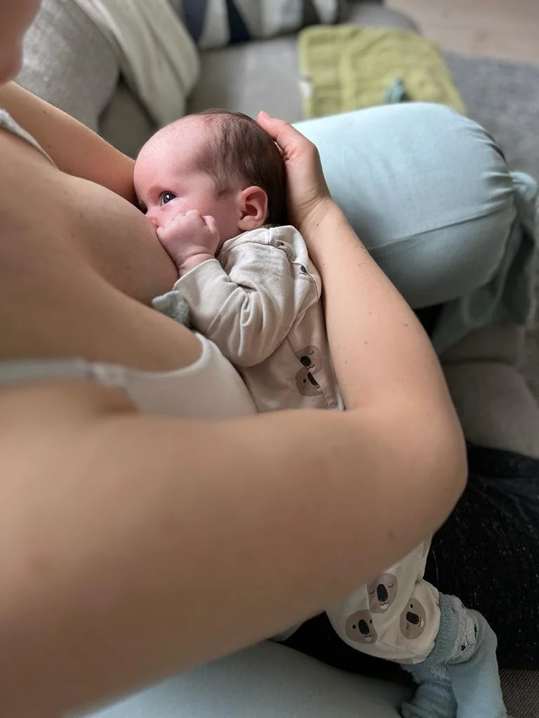 A baby breastfeeding, lying on an adult's arm with eyes open, on a sofa in a cozy living room.