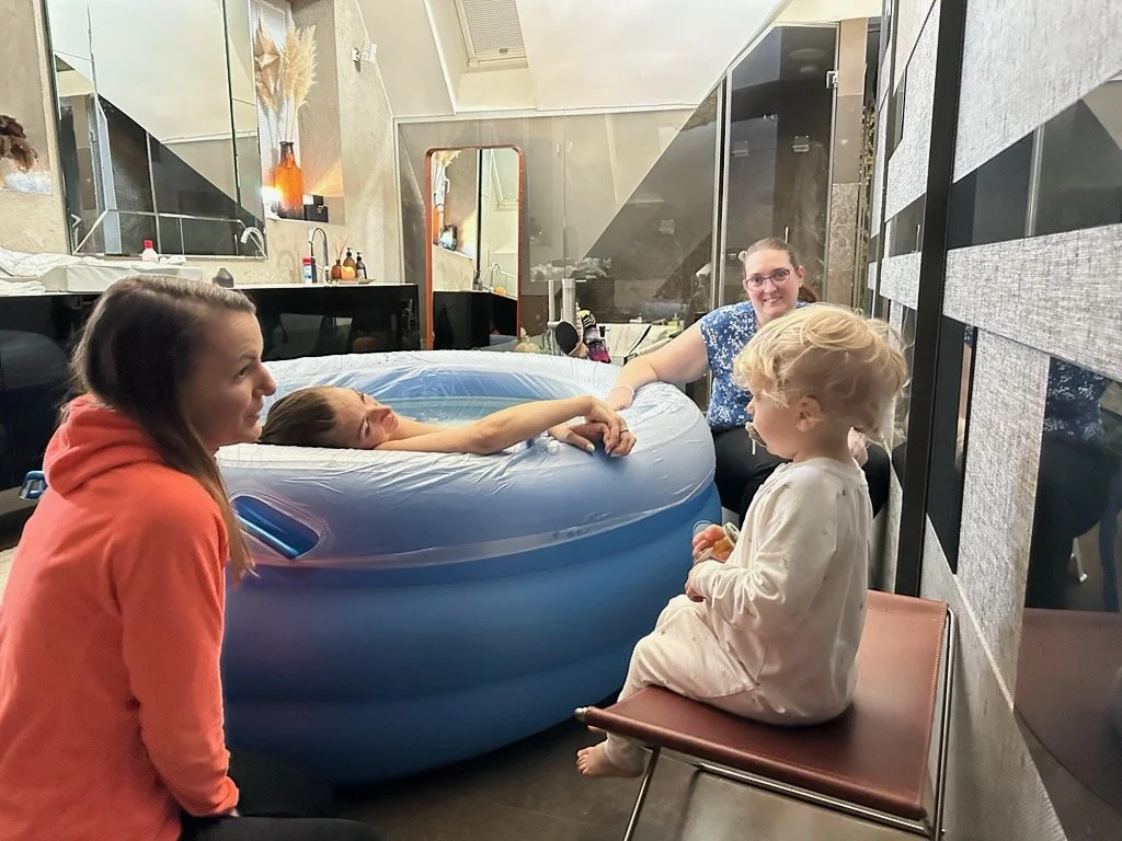 Four people gathered around an inflatable pool inside a room; one girl is floating and holding hands with a woman, while a young girl sits on a chair nearby, and another girl in an orange hoodie watches.