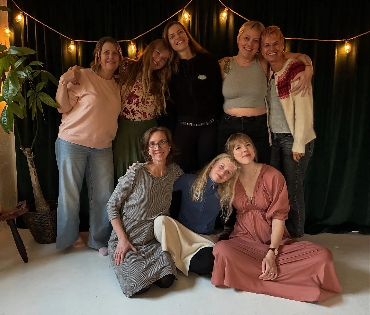 Group of eight women posing together indoors, some standing and some sitting, with string lights and a dark curtain in the background.