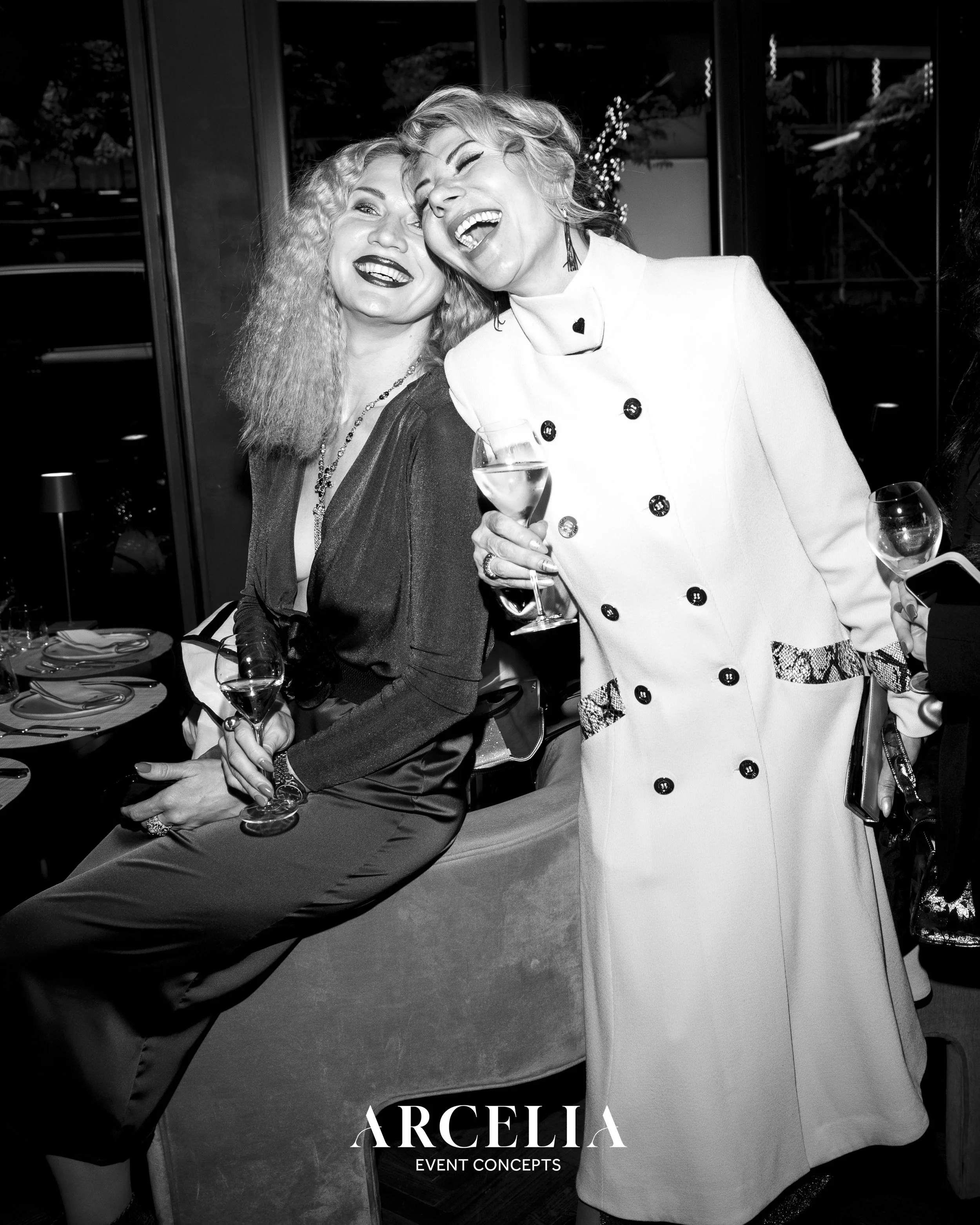 Zwei lachende Frauen beim festlichen Anlass mit Sektgläsern in der Hand, im Hintergrund dekorative Lichter und ein festlich gedeckter Tisch, in schwarz-weiß fotografiert.