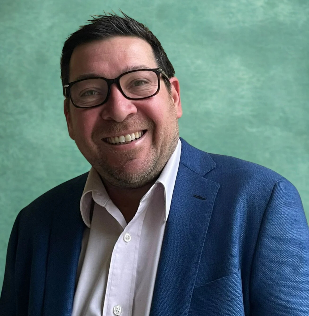 A smiling man with glasses and a beard wearing a blue jacket and pink shirt, standing against a green background.