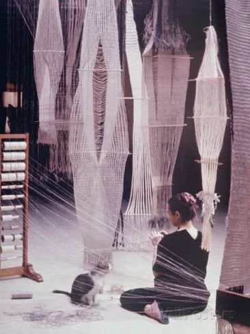 Woman seated among hanging woven textile panels and thread-filled weaving structures in a textile studio