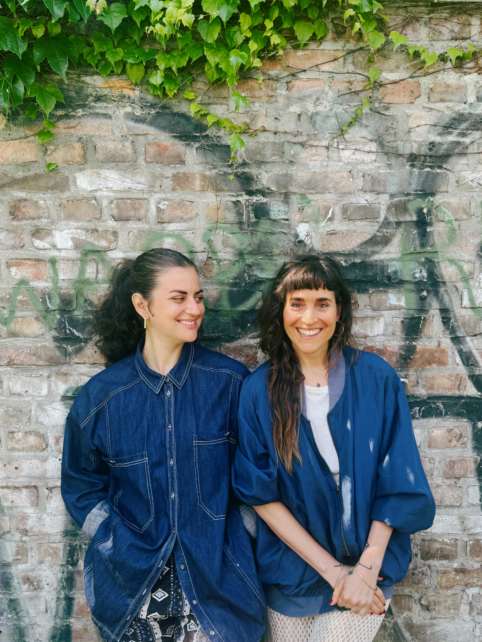 Two women smiling and standing against a graffiti-covered brick wall with green ivy growing overhead.