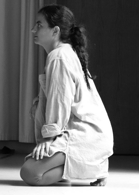Black-and-white side-profile photograph of a woman kneeling on a stage, wearing a loose white shirt and braided ponytail.
