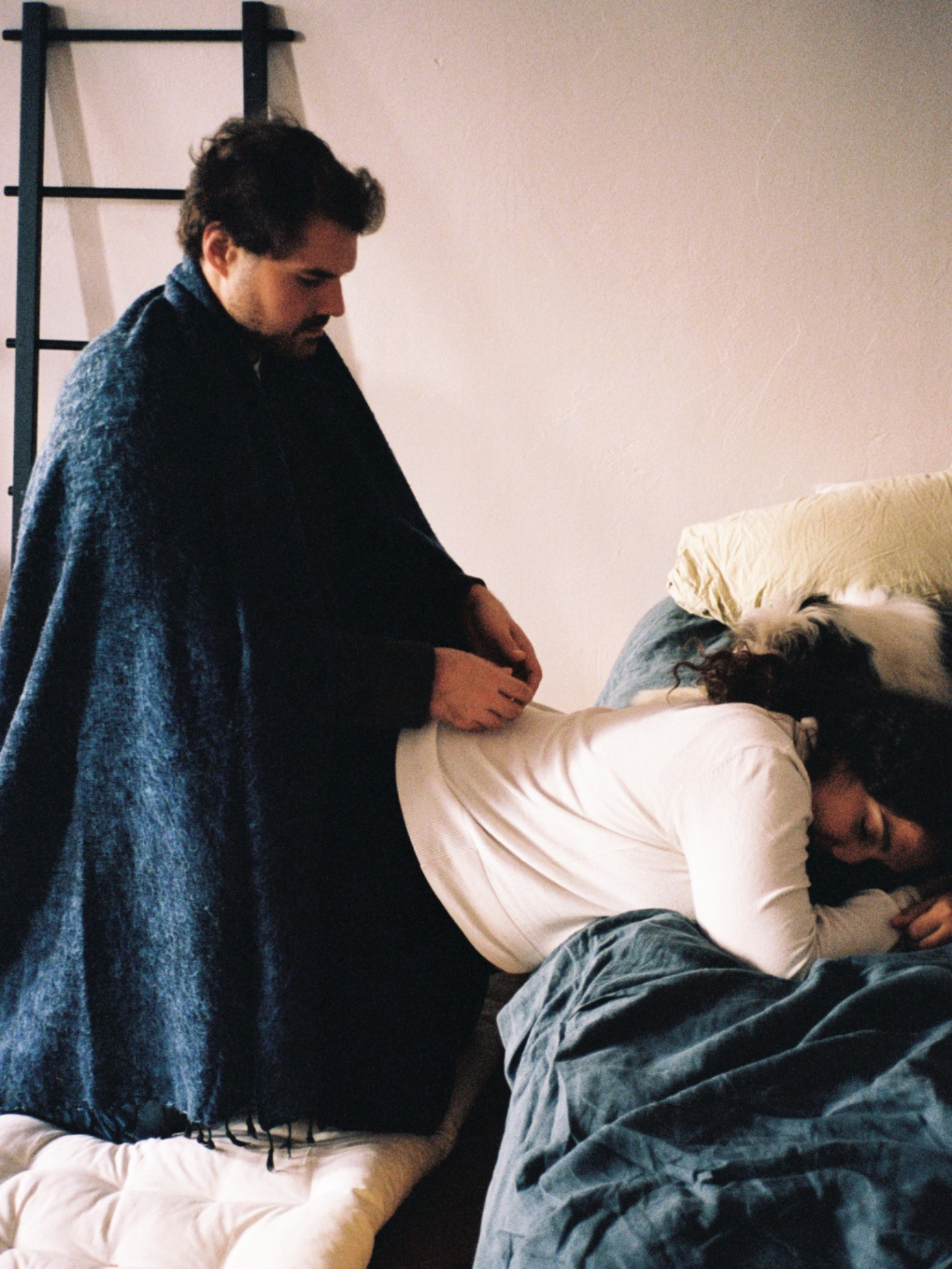 A man with dark hair and a beard helping a woman with dark curly hair lying on a bed, with a pillow and blanket. The man is wearing a dark sweater, and the woman is wearing a white top and denim jeans.