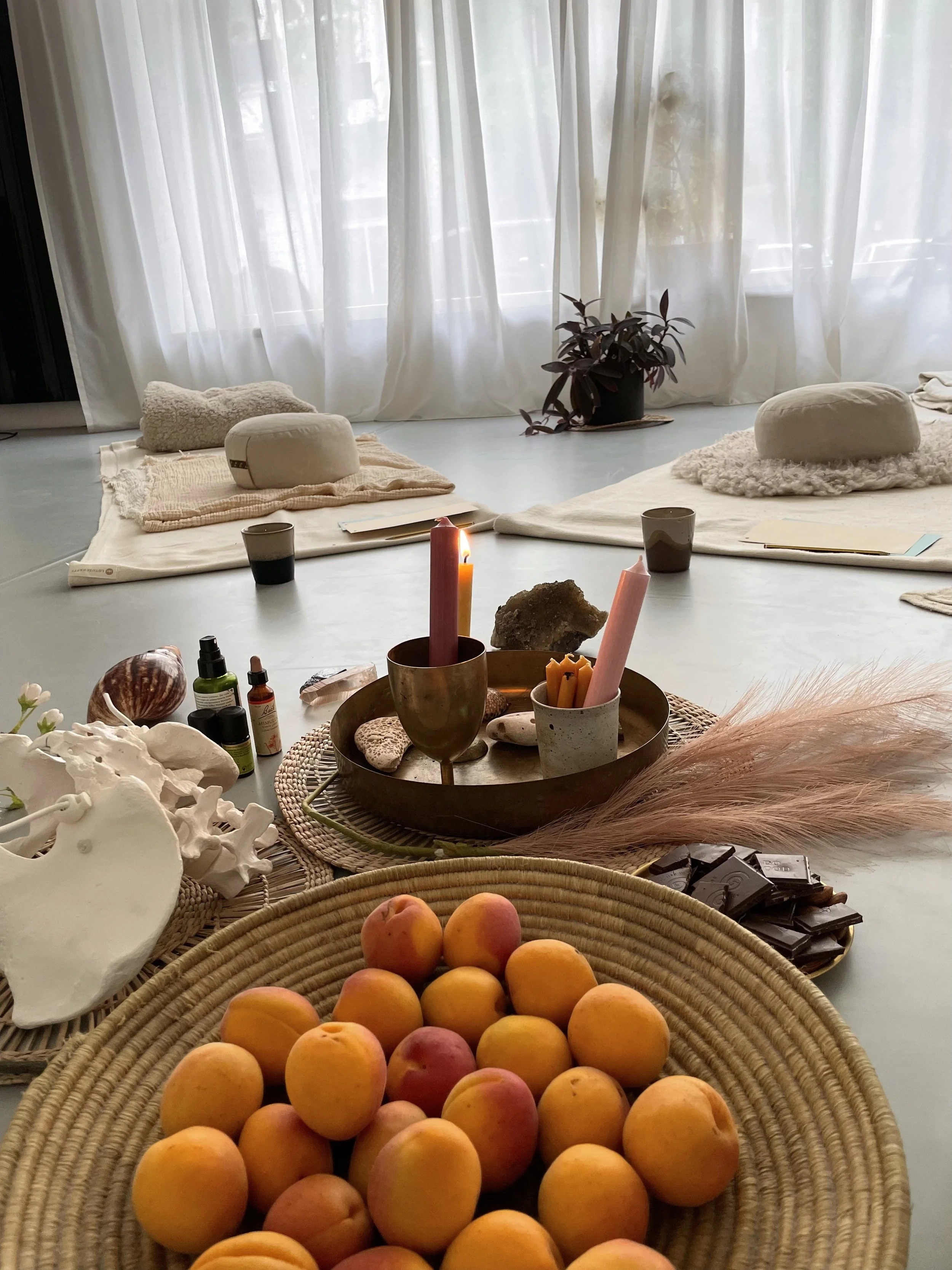 Cozy wellness setting with sheer curtains, floor cushions, candles, crystals, essential oils, flowers, dark chocolate, and a basket of apricots.