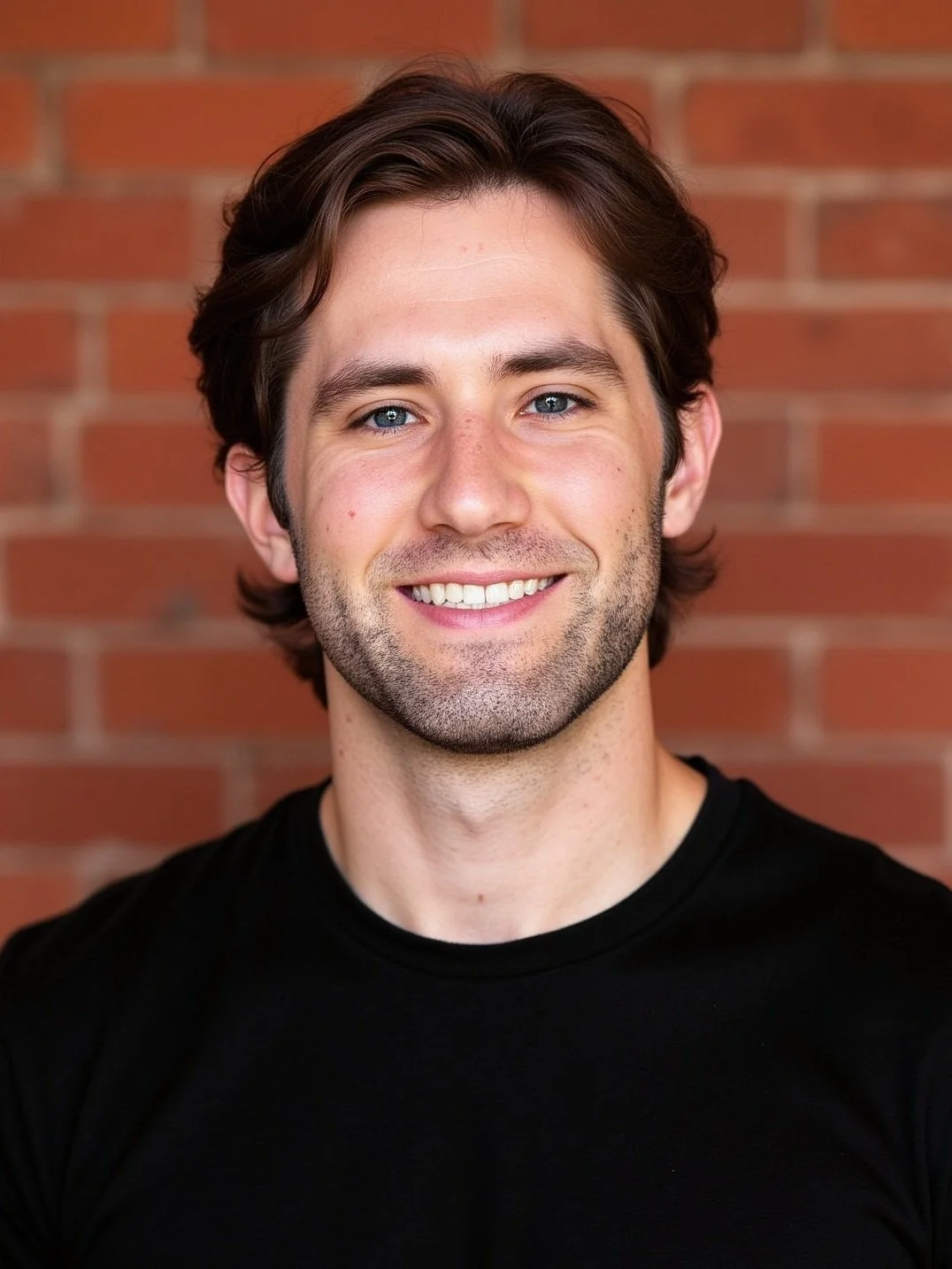 A young man with brown hair, blue eyes, and light skin smiling against a brick wall background.