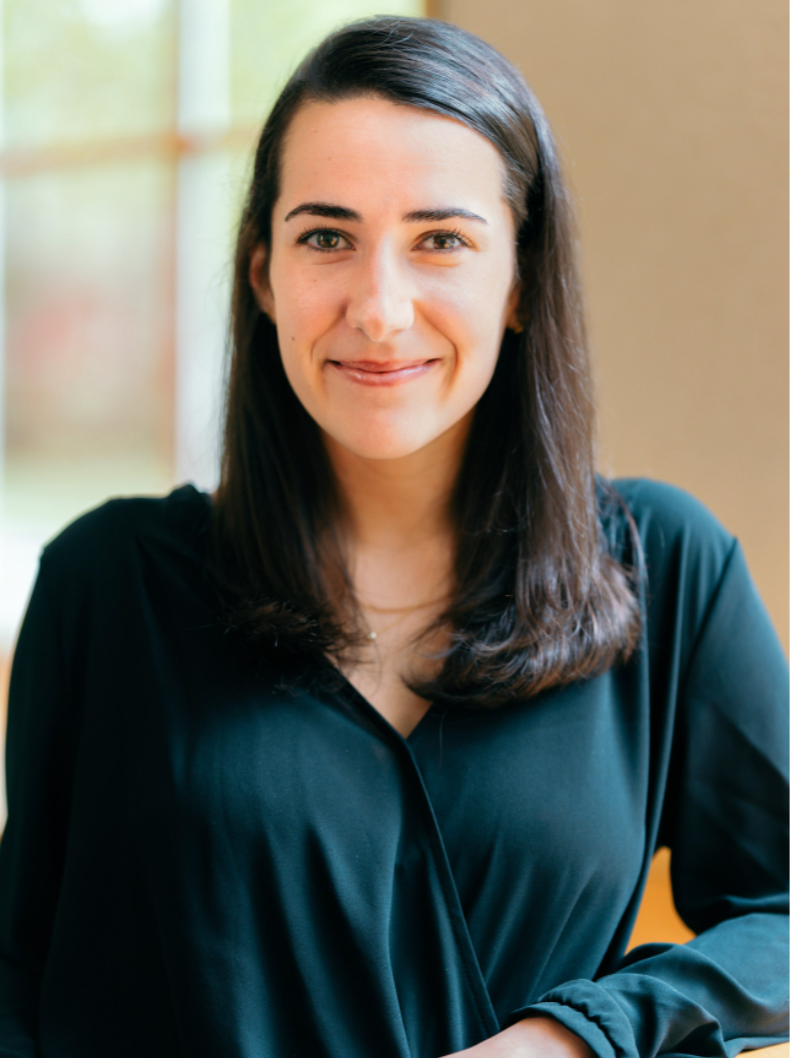A woman with long brown hair and fair skin smiling at the camera, wearing a black shirt inside a room with natural light and a blurred background.