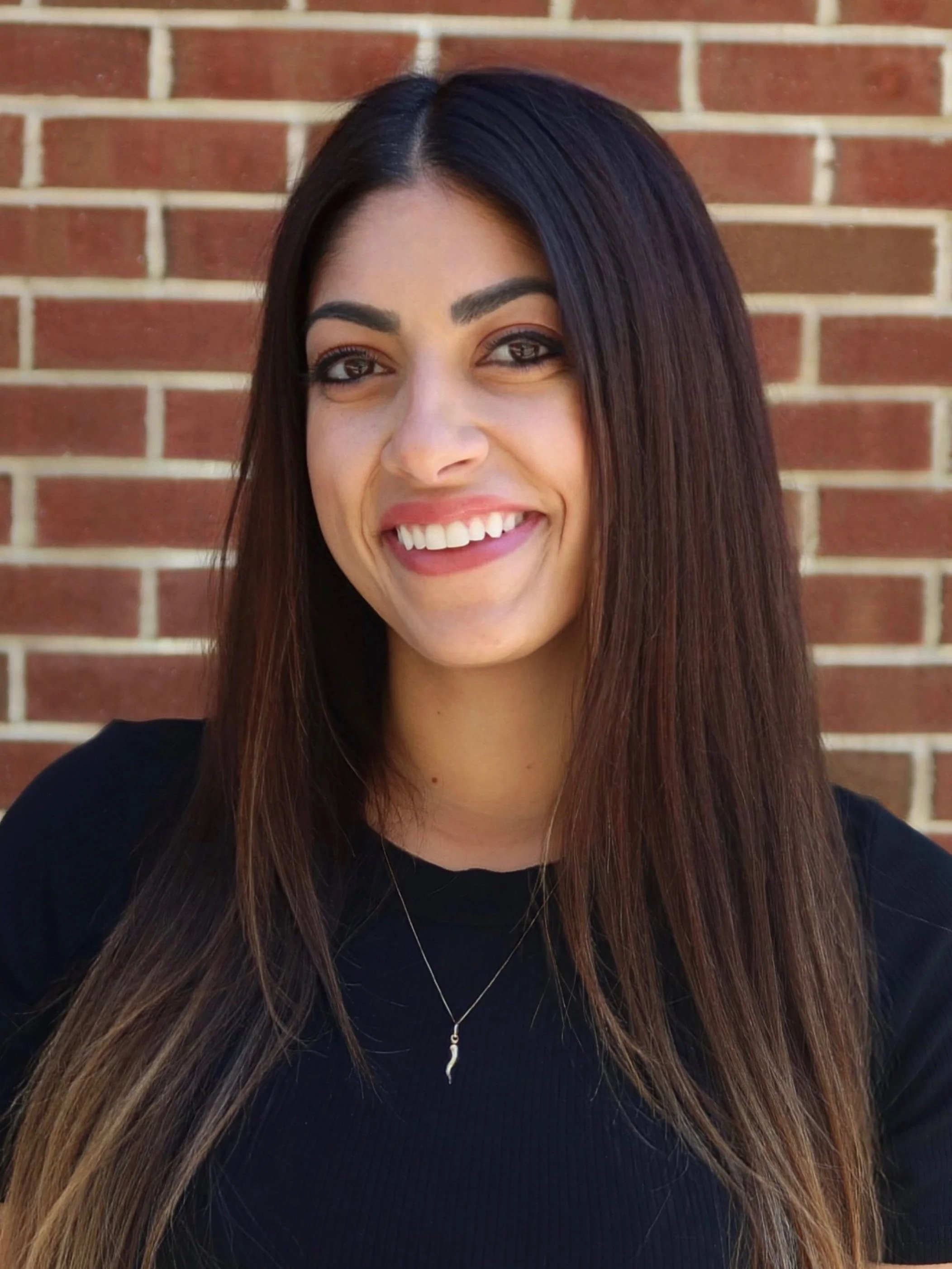 Smiling woman with long brown hair and dark eyes standing in front of a brick wall.