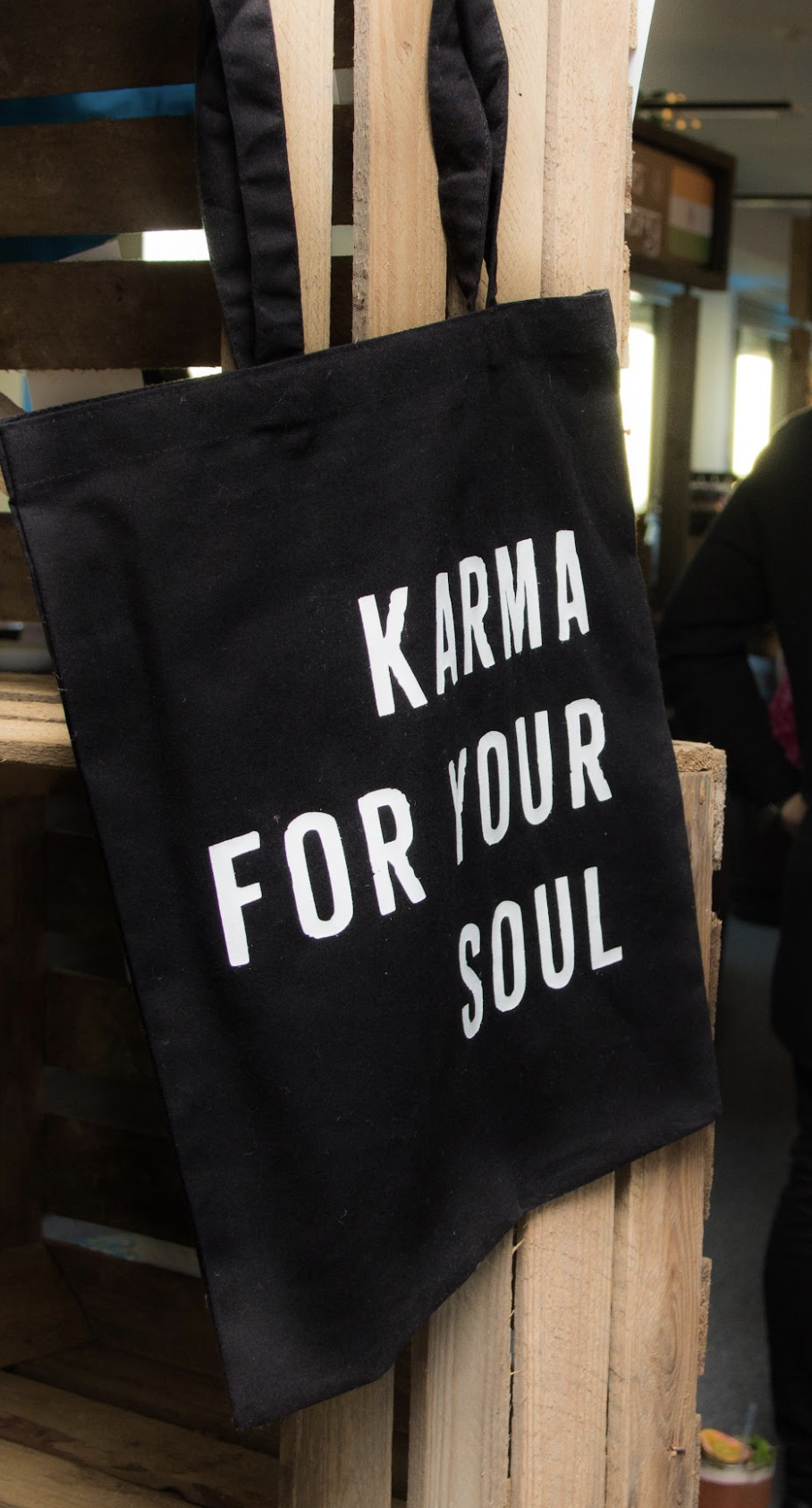 A black tote bag hanging on a wooden pallet with white text that reads 'KARMA FOR YOUR SOUL'.