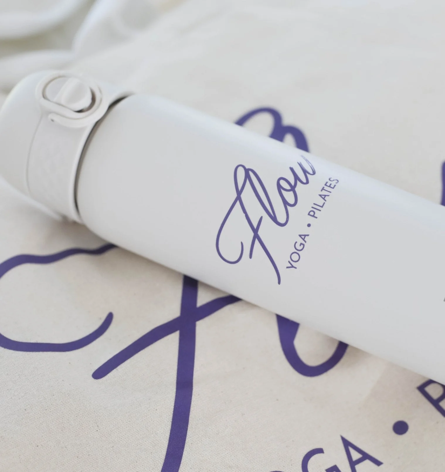 Close-up of a white cylindrical object with a purple logo that says "Flow" and the words "YOGA. PILATES" underneath, placed on a fabric surface with purple script text.