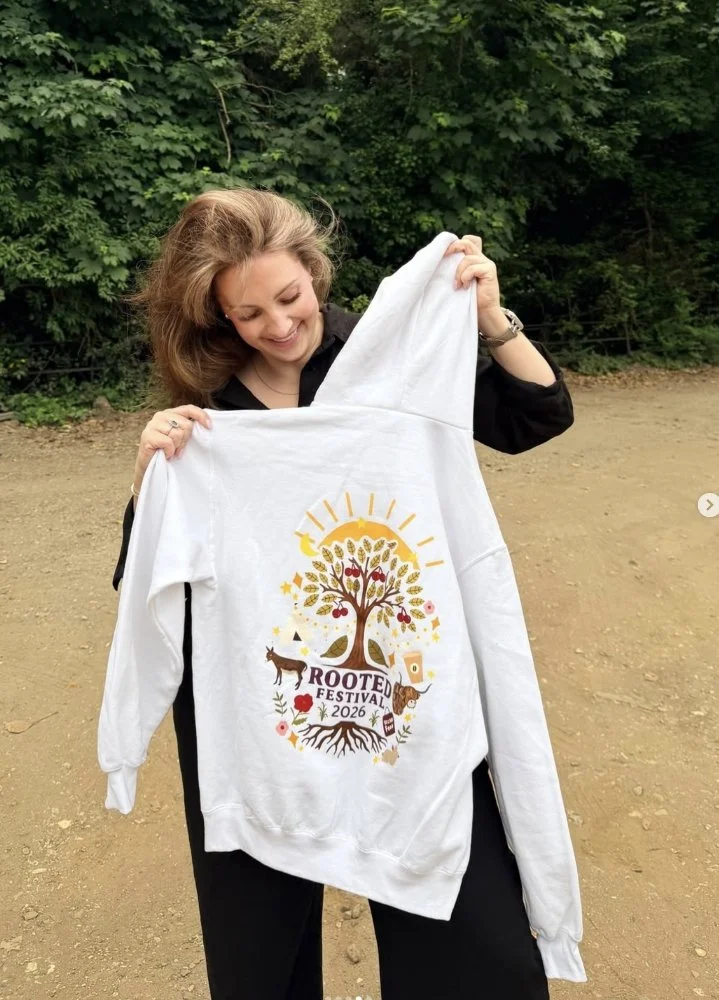 Woman smiling outdoors, holding a white hoodie with a colorful design of a tree, animals, and the text 'Rooted Festival 2026'.