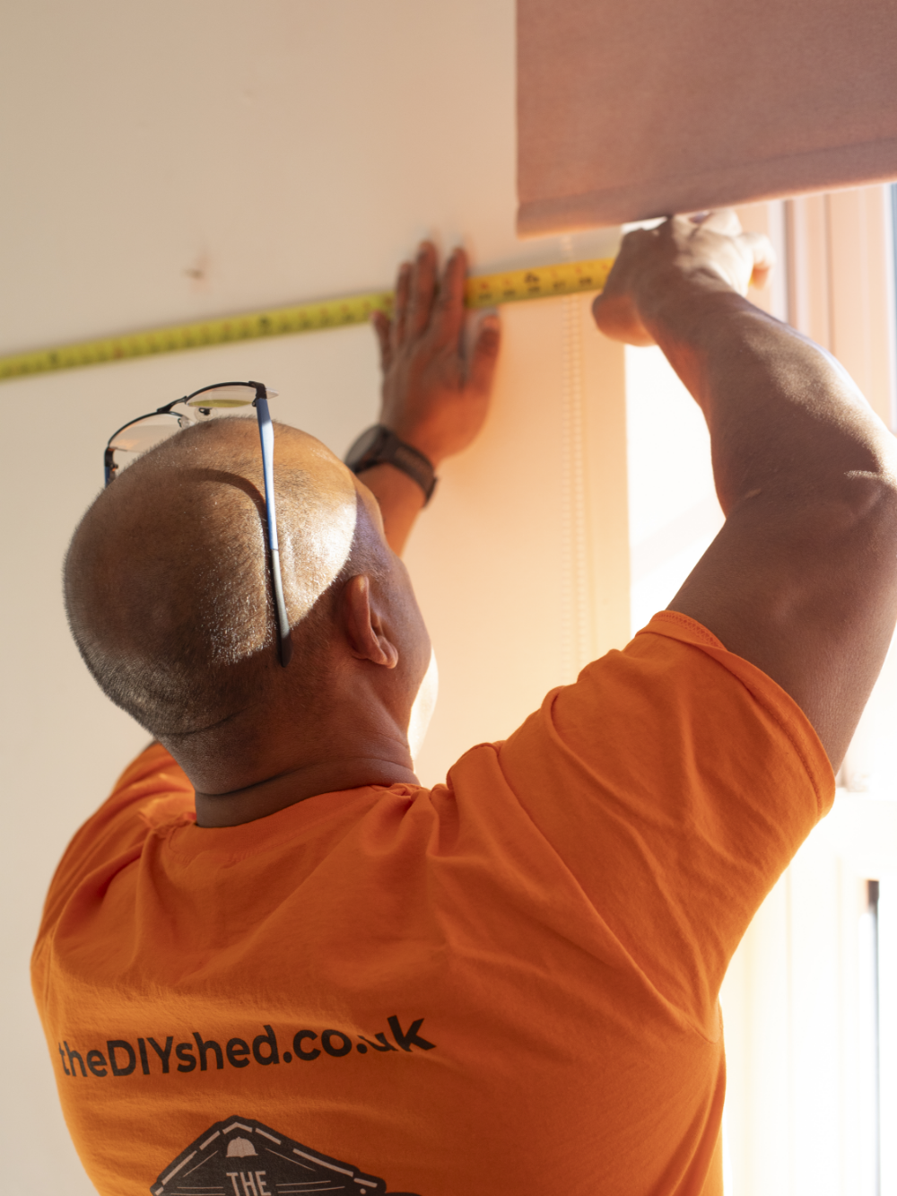 A man wearing sunglasses and an orange shirt measuring a wall with a yellow tape measure near a window.