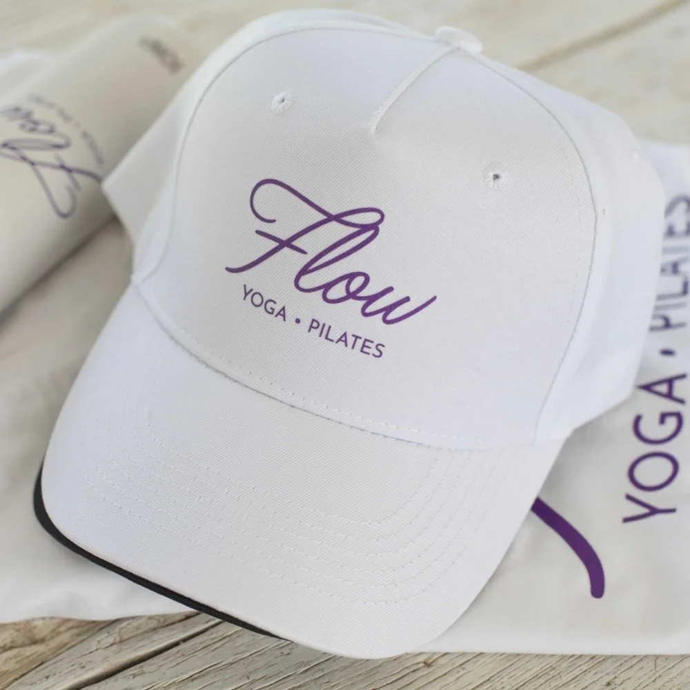 White baseball cap with purple text that reads "Flow" and smaller text below saying "Yoga . Pilates". The cap is resting on a white cloth with similar text, placed on a wooden surface.