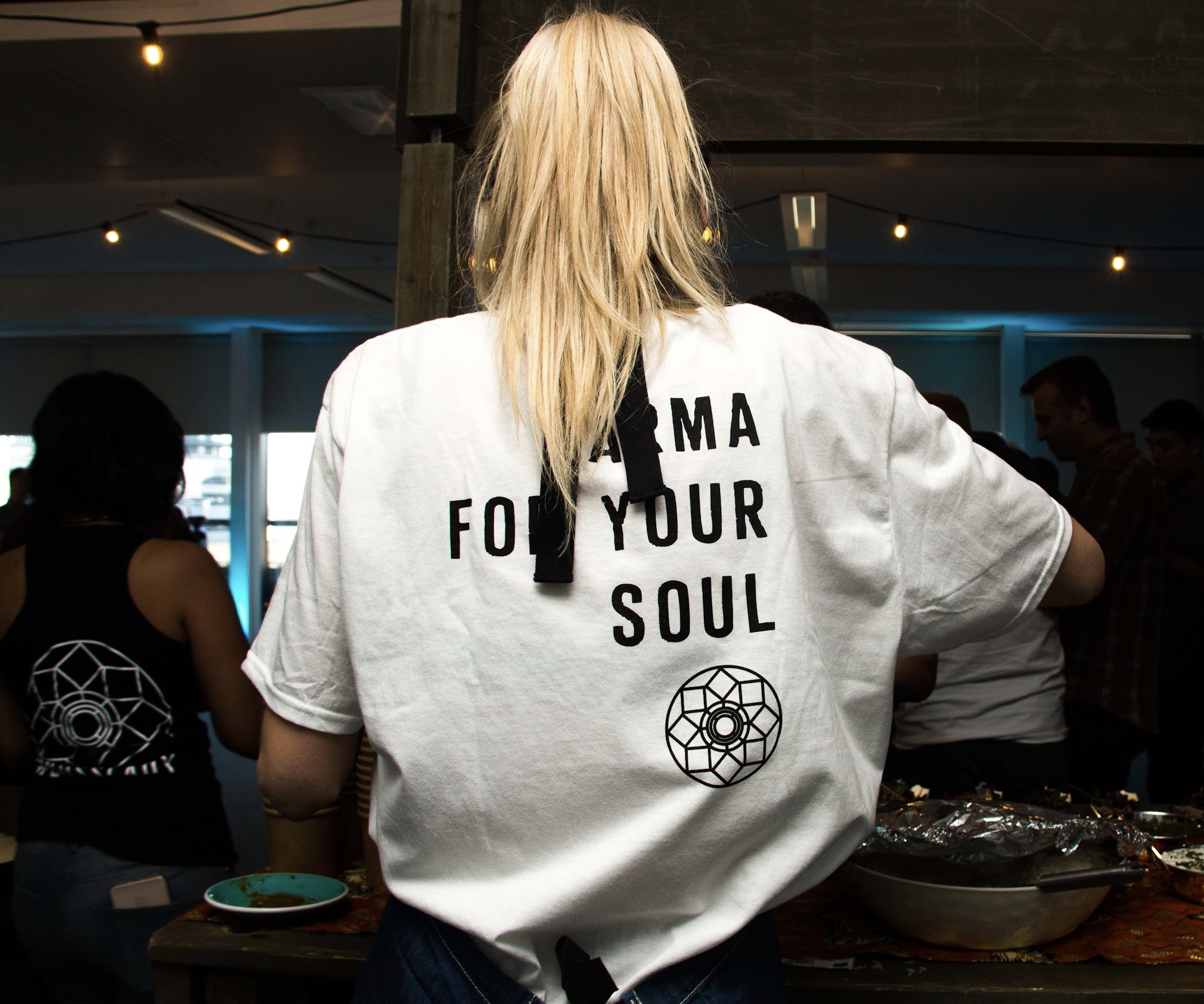 Back view of a woman with blonde hair wearing a white T-shirt with black text, in a room with several people and a table with food and drinks.