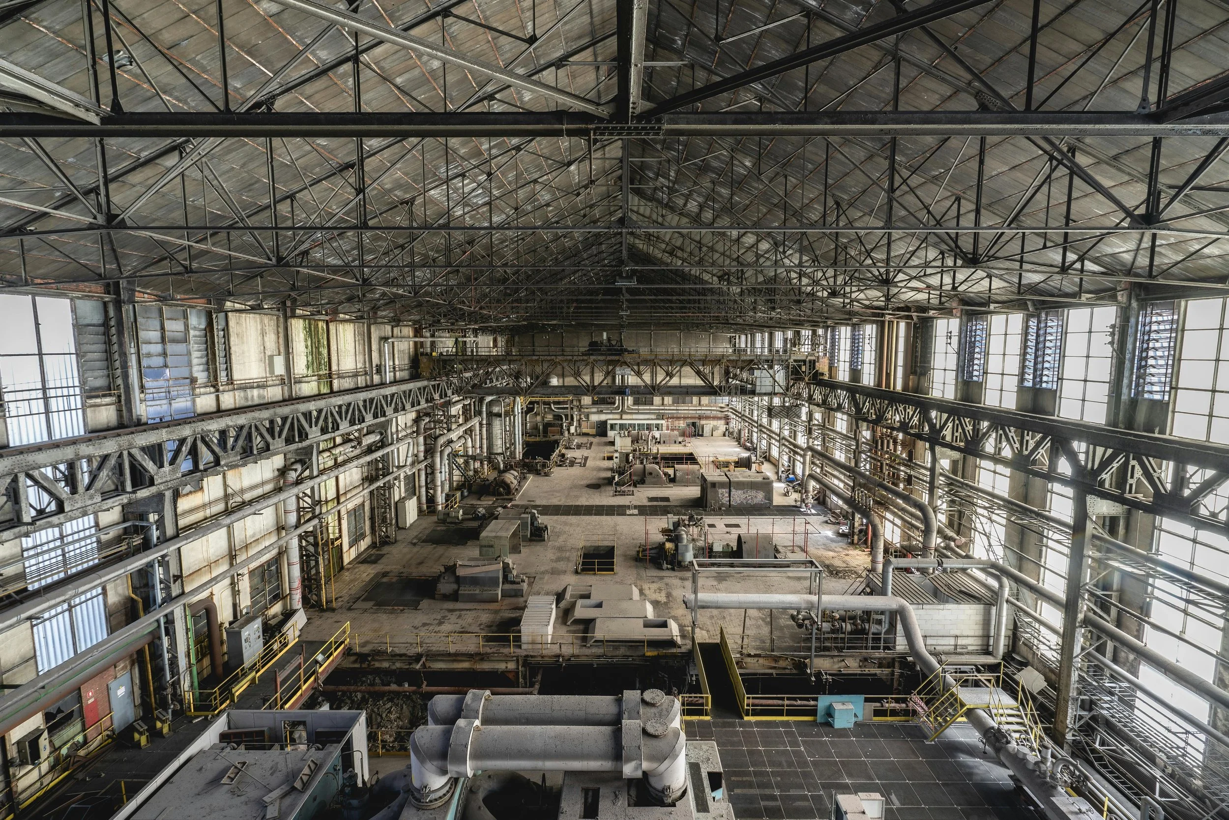 Interior of an abandoned industrial factory or warehouse with large empty space, exposed metal beams, pipes, and windows allowing natural light.