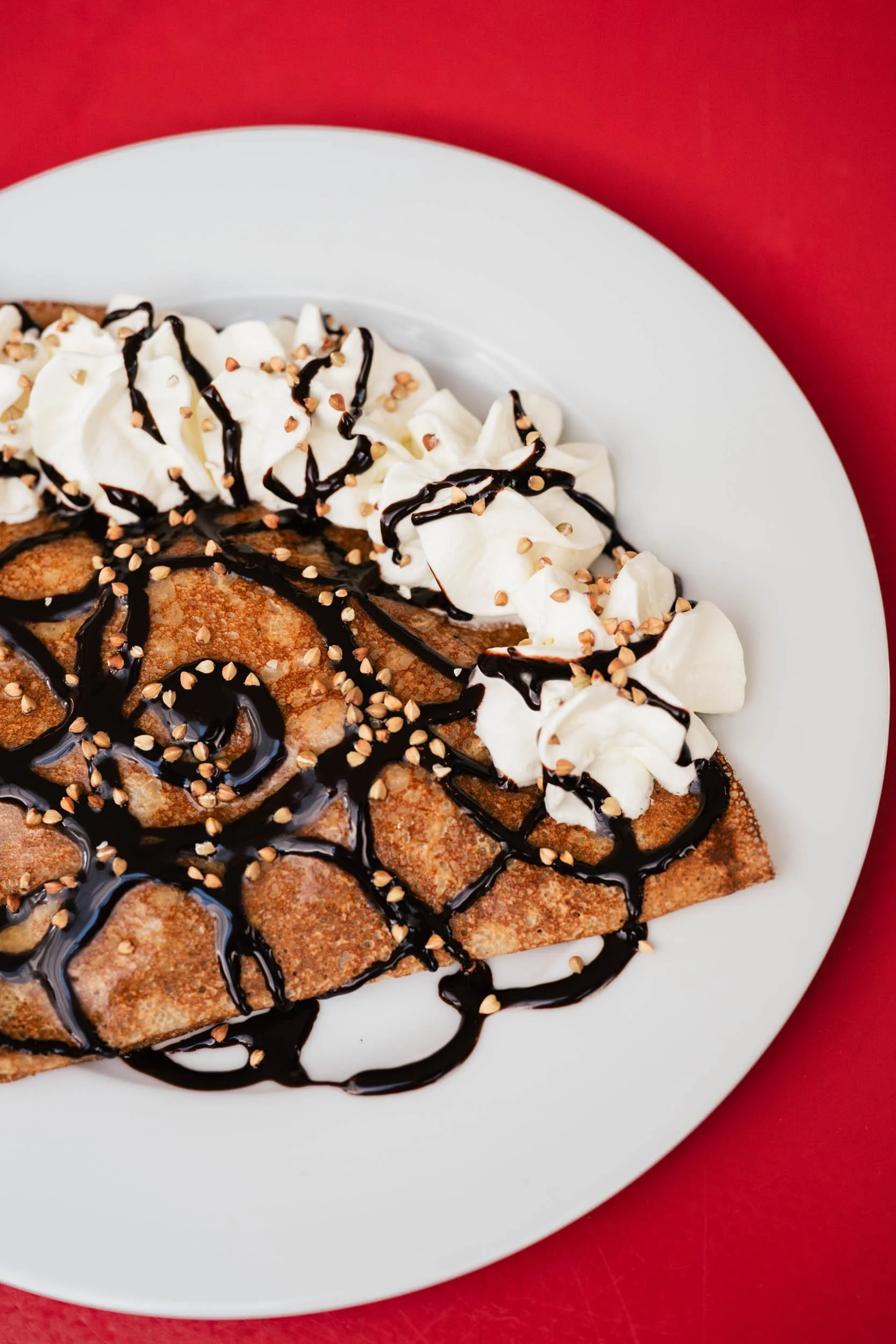 Crêpe garni de chantilly, chocolat et noix, présenté sur une assiette blanche contre un fond rouge.