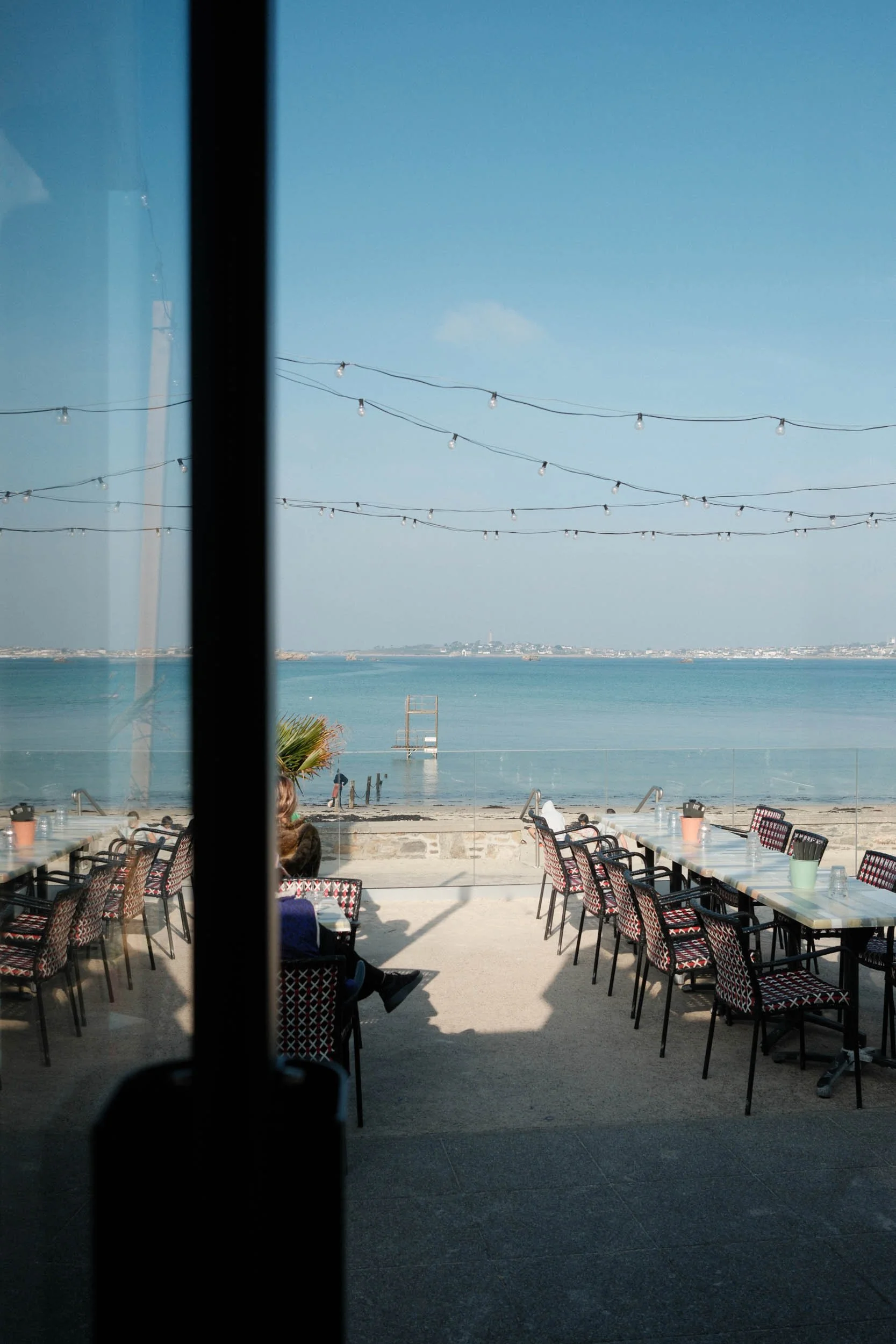 Vue d'une terrasse de café avec des tables et des chaises, regardant vers la mer avec un ciel bleu clair, des guirlandes lumineuses suspendues au-dessus, à travers une porte vitrée.