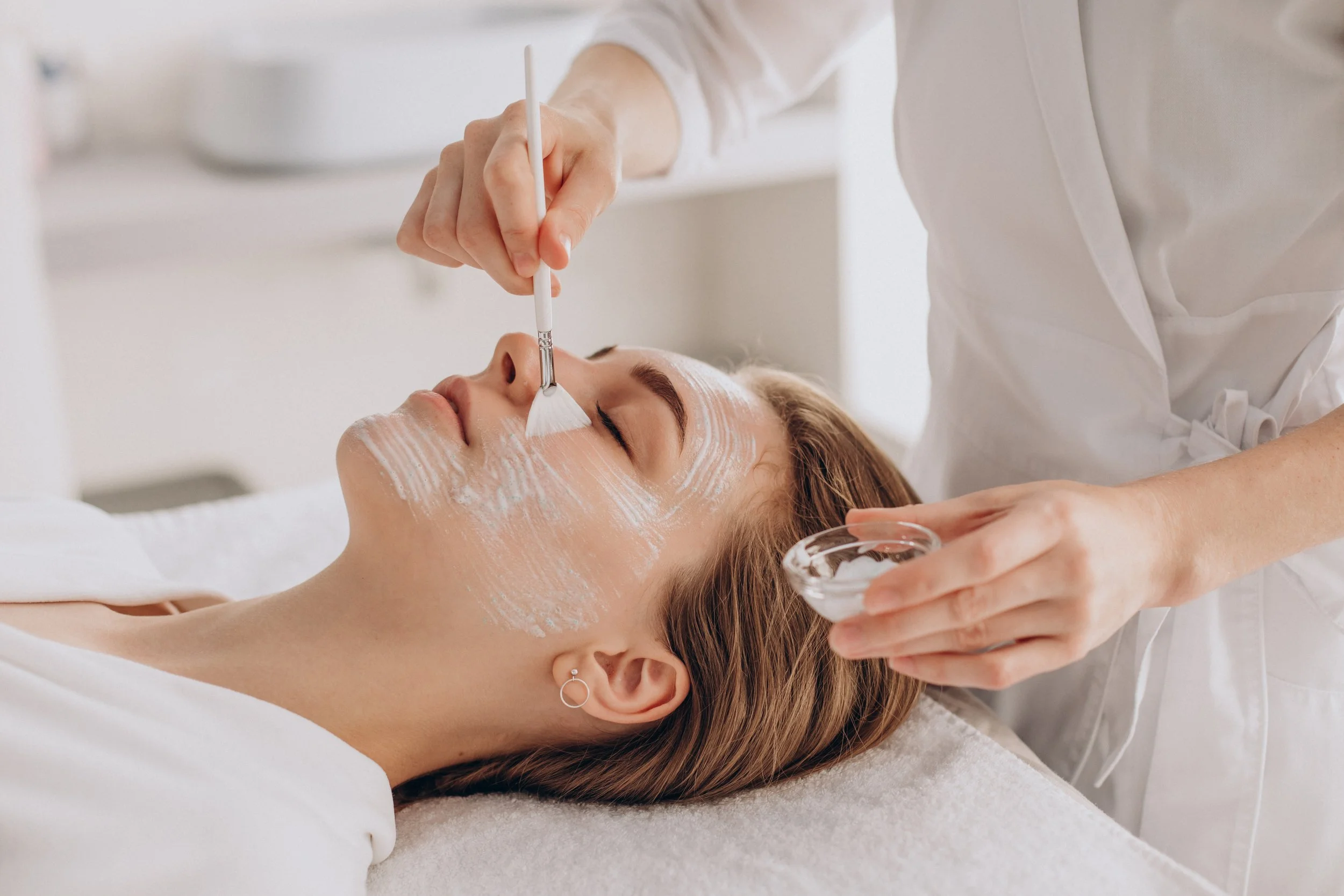 A woman receiving a facial treatment from a skincare professional at a spa or clinic, with milky white substance applied to her face.