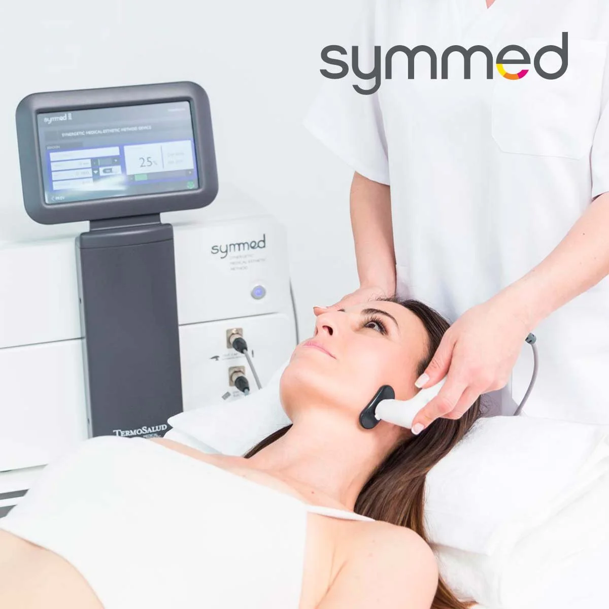 A woman receiving a facial treatment with a device on her cheek in a medical spa or clinic while a technician operates a machine with a screen.