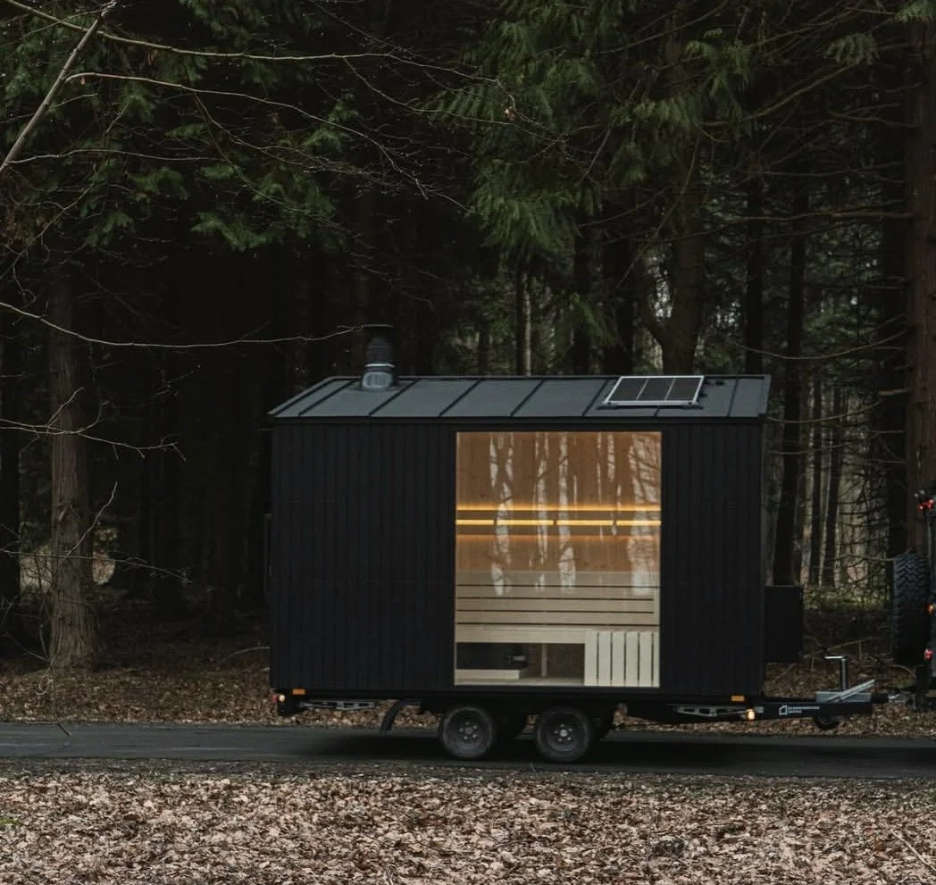 A black tiny house trailer with a large window, surrounded by tall trees in a forest setting.