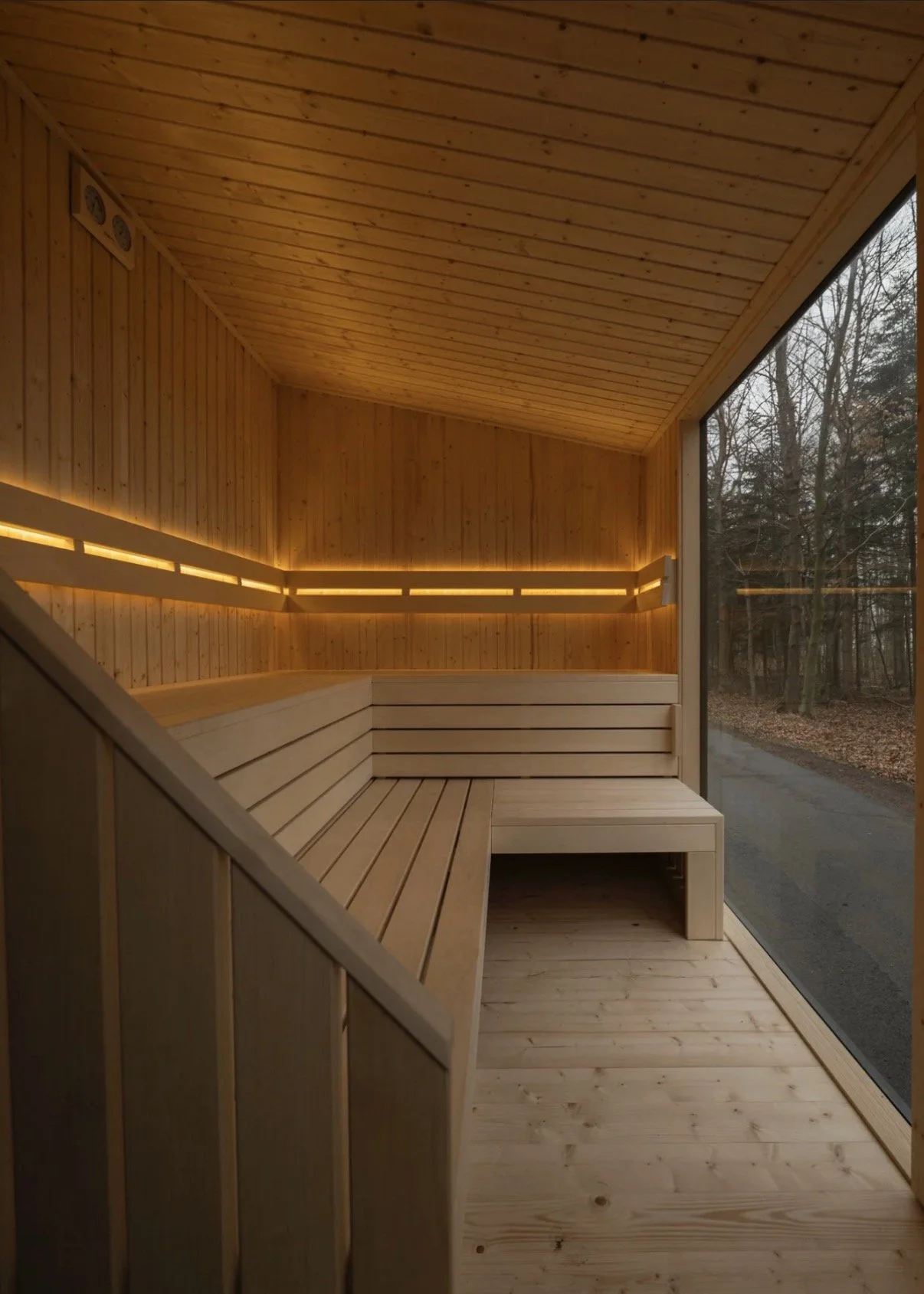 Interior of a modern wooden sauna with built-in bench seating, warm lighting, and a large window showing an outdoor wooded landscape.