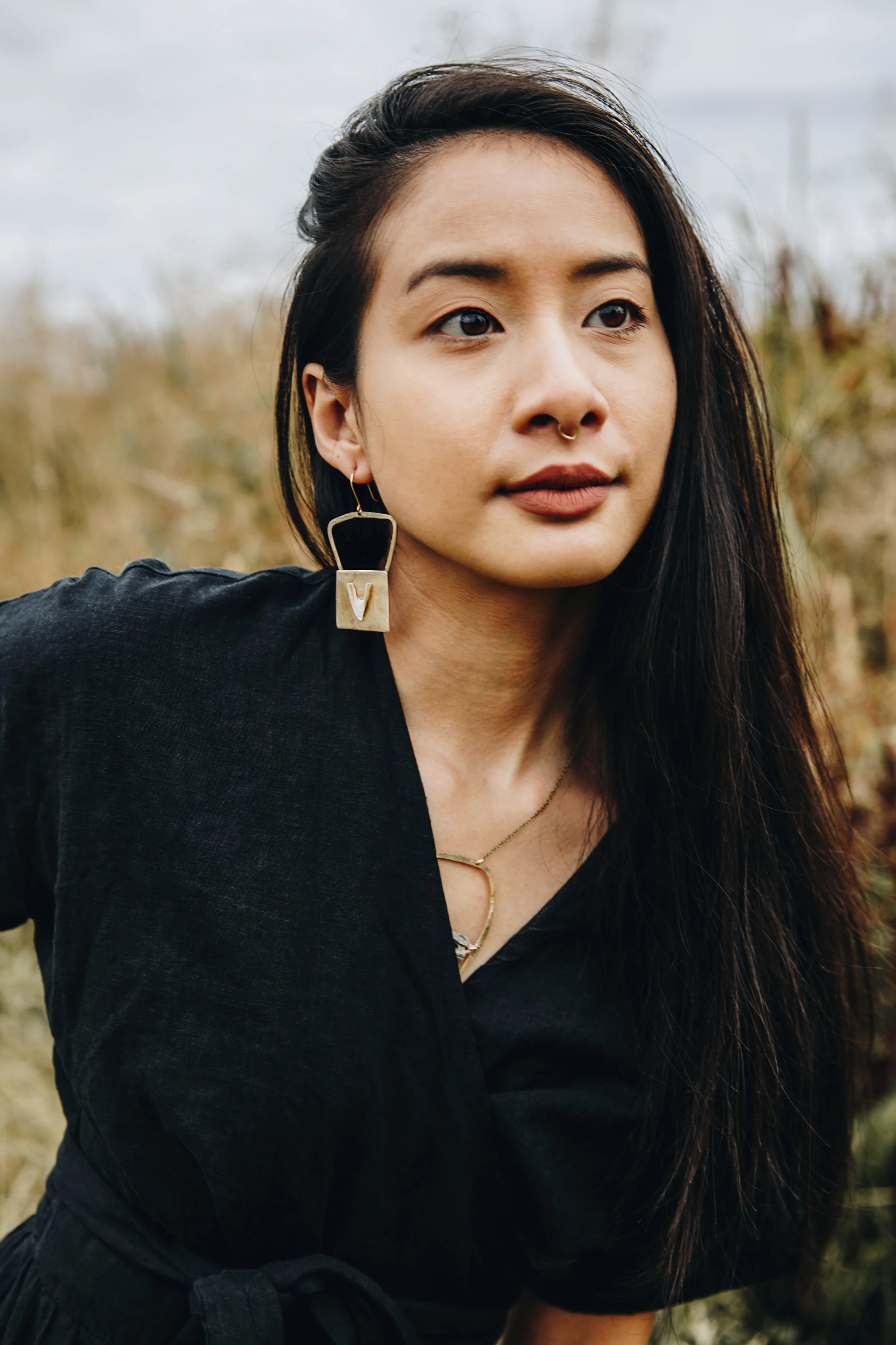 A young woman with long dark hair, wearing a black top and jewelry, standing outdoors near tall grass, with a cloudy sky in the background.
