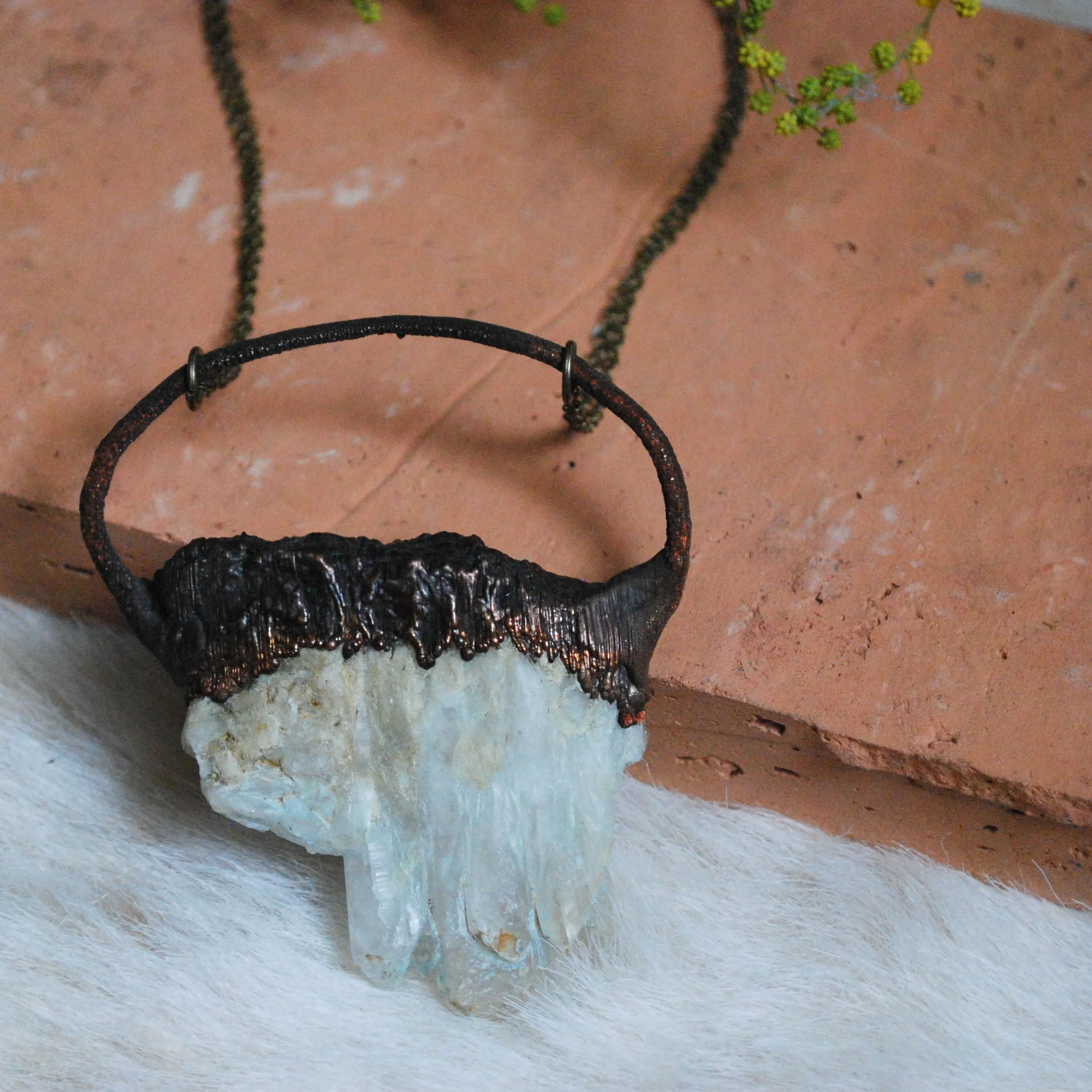 Necklace with a large pendants made of carved stone on electroformed copper, hanging from a black cord, placed on a soft white surface with a brick wall in the background.