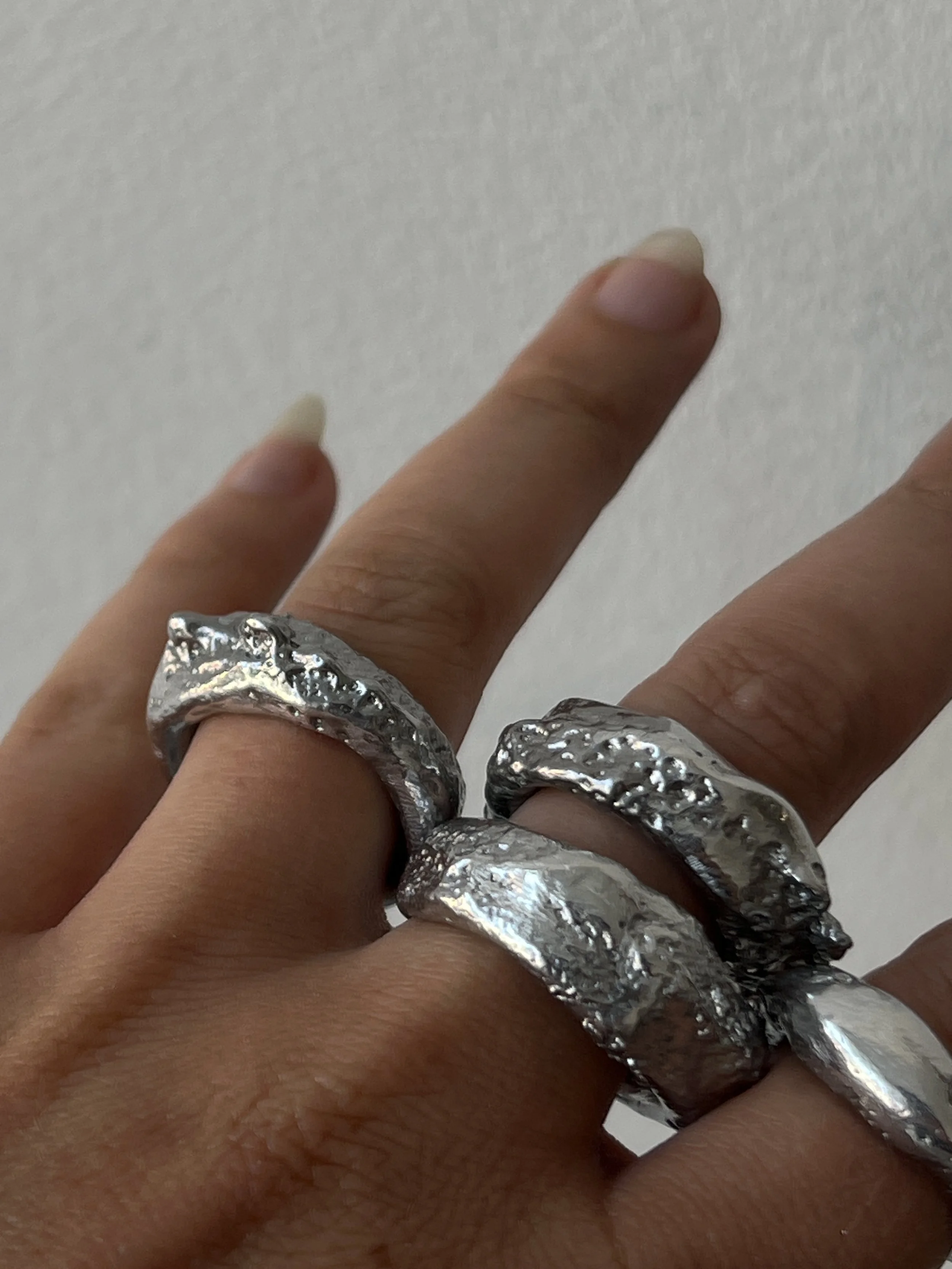 Close-up of a hand with three silver rings on the fingers, each with a rugged, textured design, against a plain background.