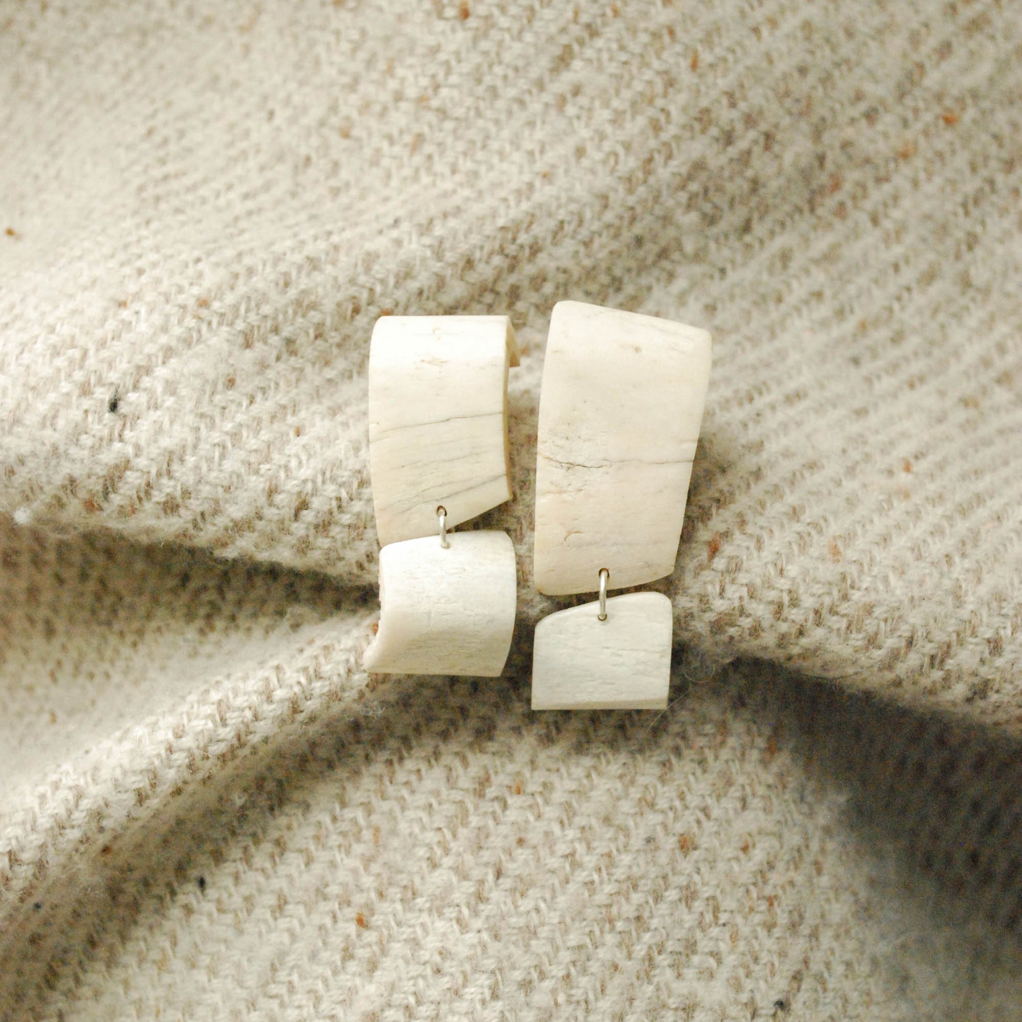 A pair of white earrings made from bone with curved rectangular and square shapes resting on a textured beige fabric background.