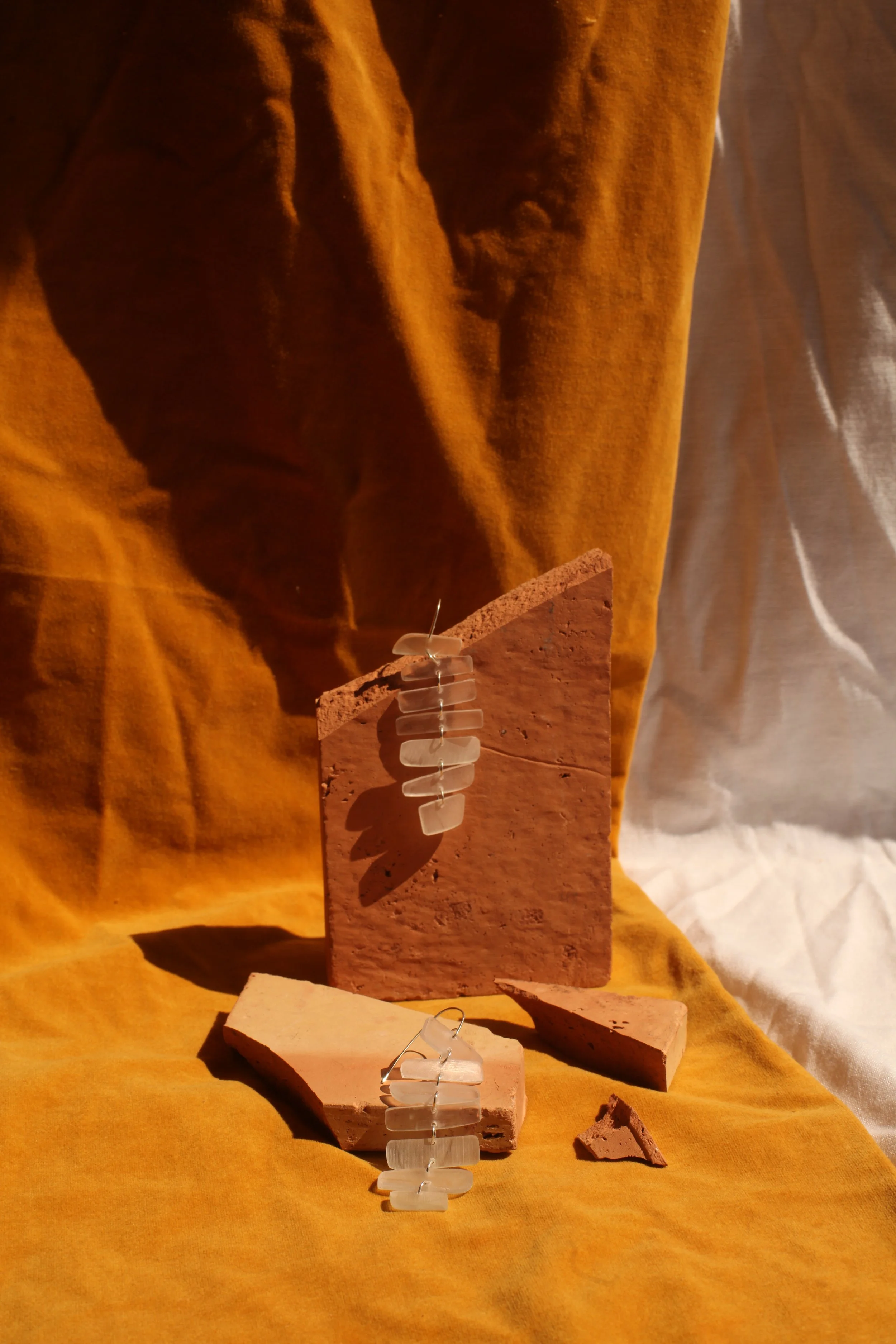 Artistic still life featuring a pair of earrings made of white rectangular beads cut from old glasses lenses, displayed on beige and terracotta-colored tiles with a yellow fabric background and a white curtain.