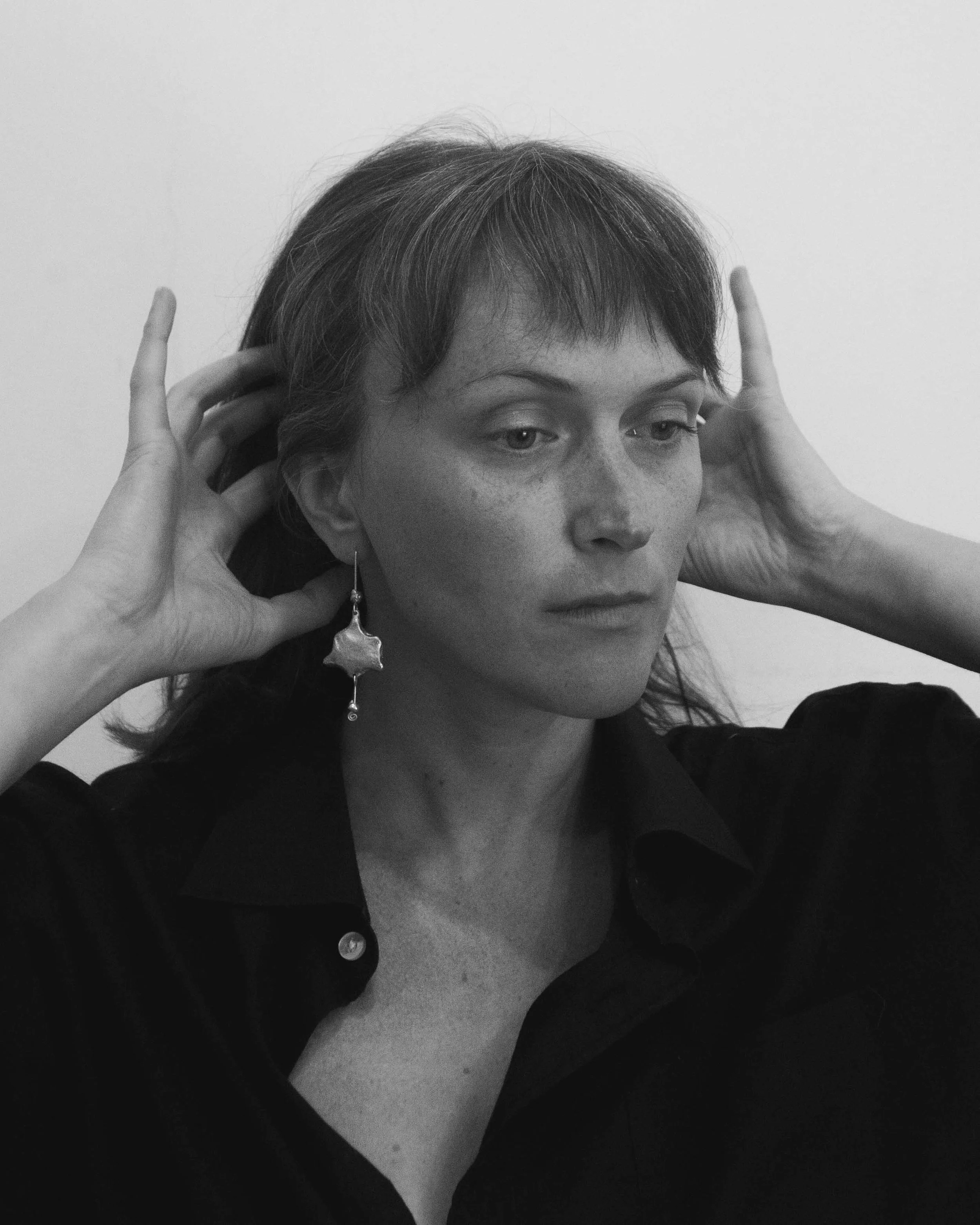 A woman with short hair wearing a black collared shirt, holding her hands near her head, with a large star-shaped earring on her left ear, looking contemplative.