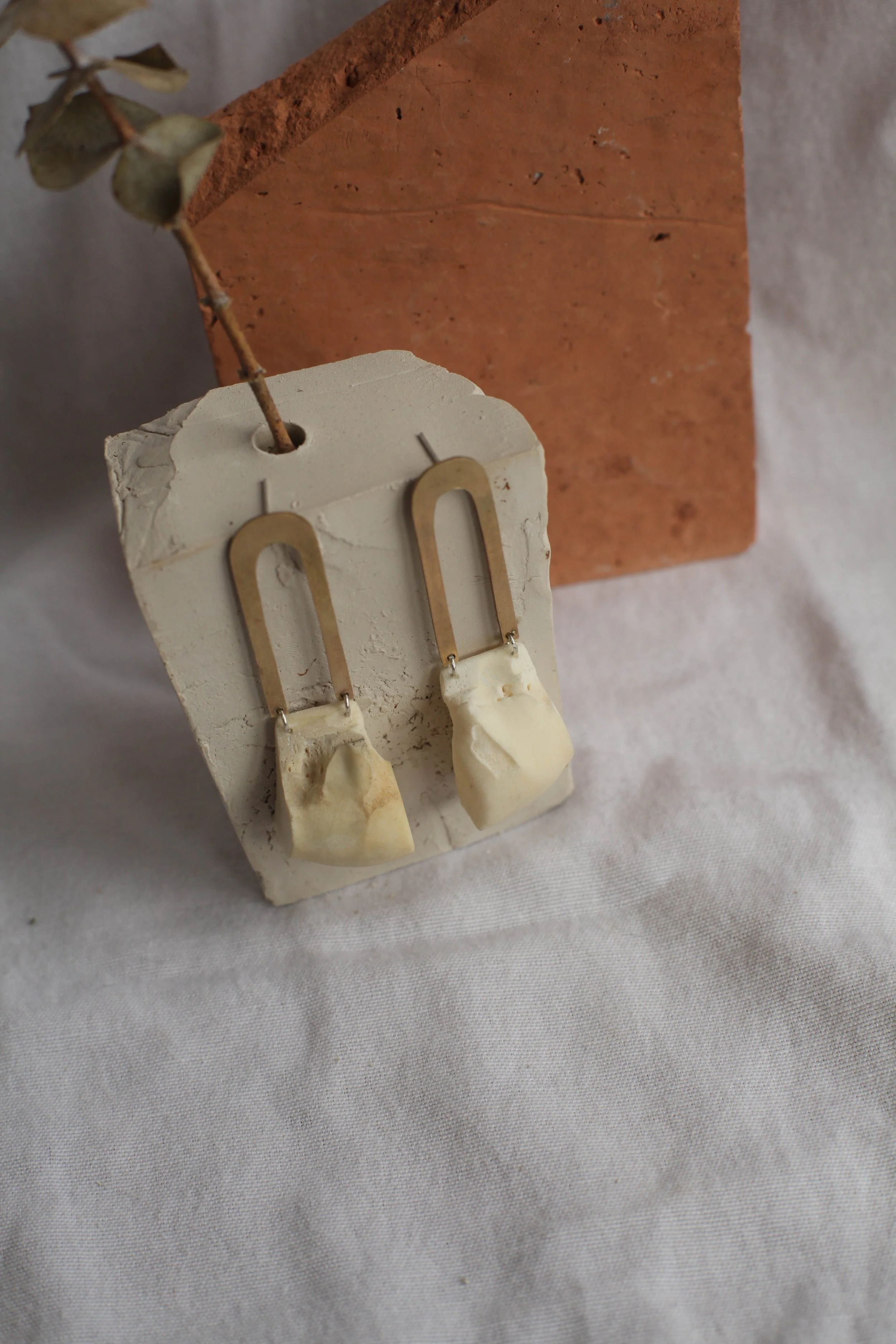 A pair of modern earrings with bone pendants and brass hooks displayed on a small white square block, with a dried plant and a brick in the background.