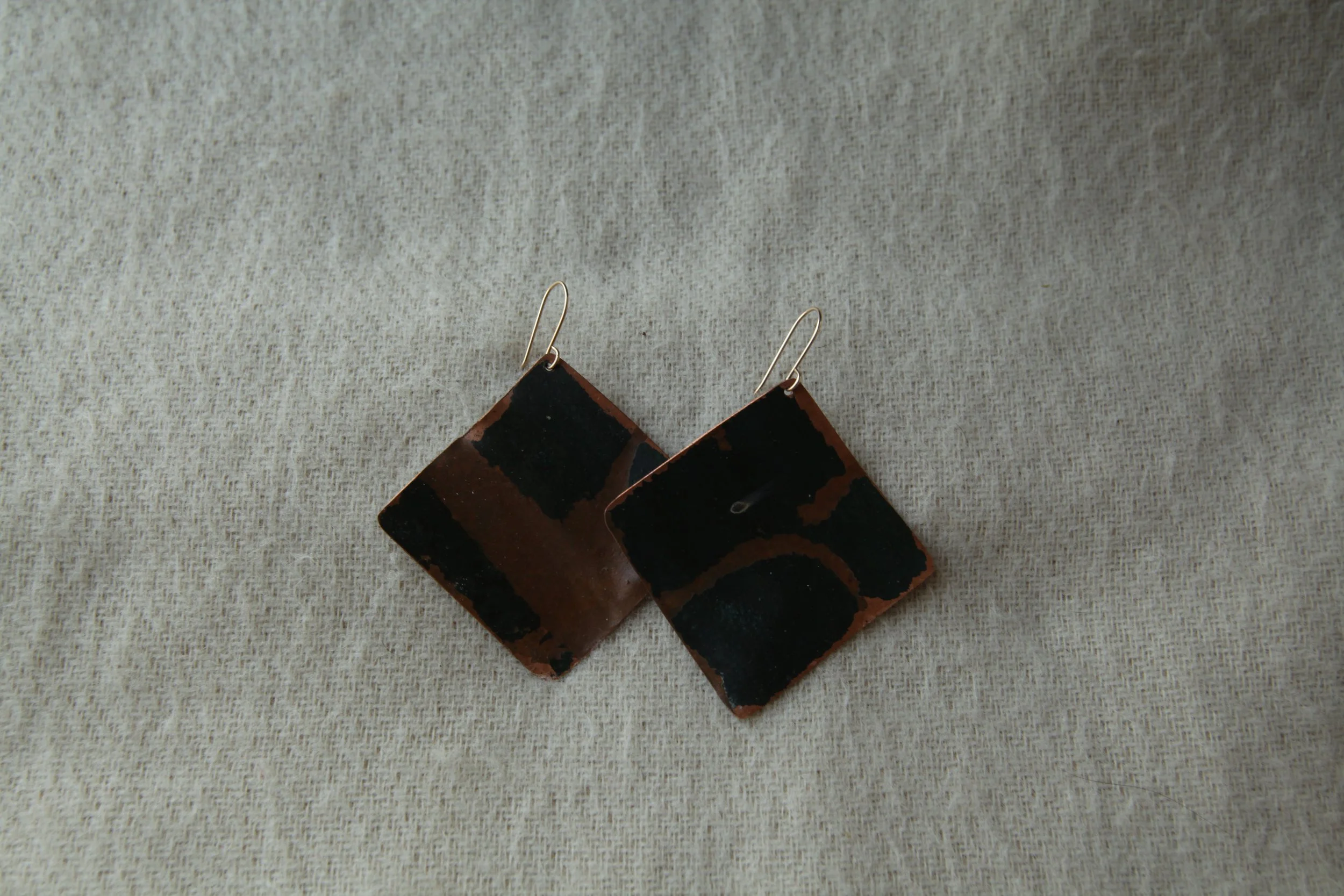 Pair of square-shaped earrings with black and brown geometric patterns on a beige fabric background.