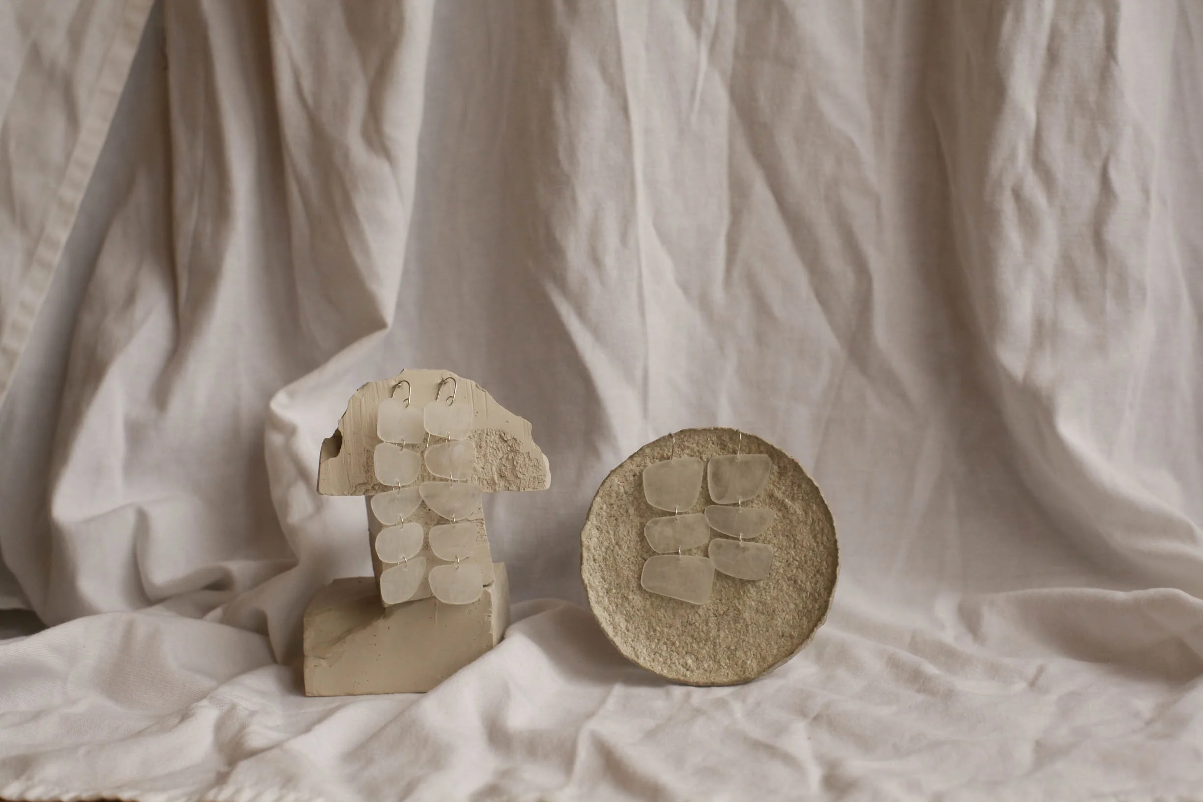 Two ceramic jewelry displays, both with earrings made from upcycled eye glass lenses, are set against a draped white fabric background.
