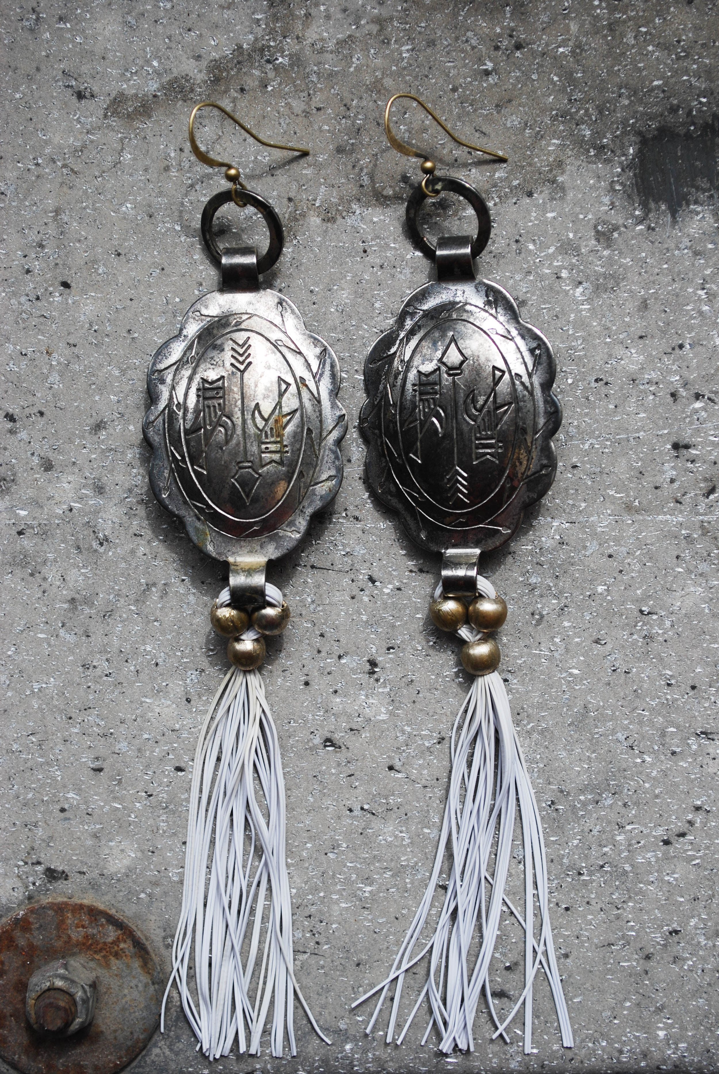 A pair of ornamental silver earrings with engraved designs, dangled with small brass balls and white tassels, placed on a rough concrete surface.