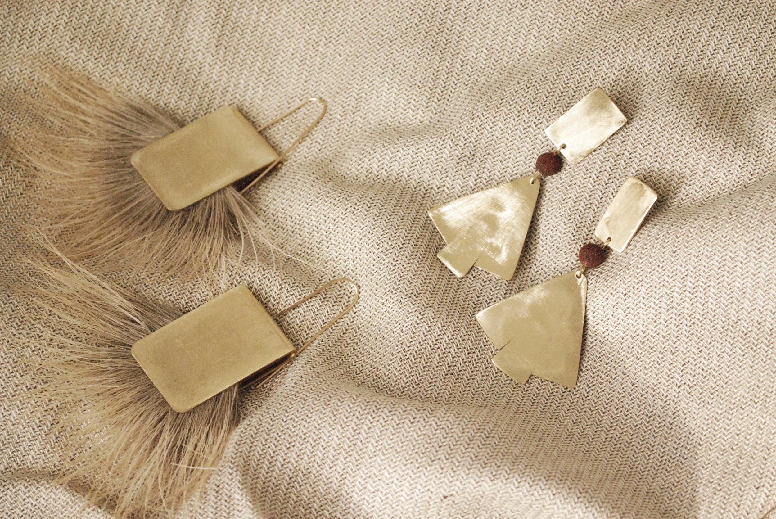 Brass fringe earrings, brass geometric earrings, and rectangular earrings with brown beads, all displayed on a textured beige fabric background.