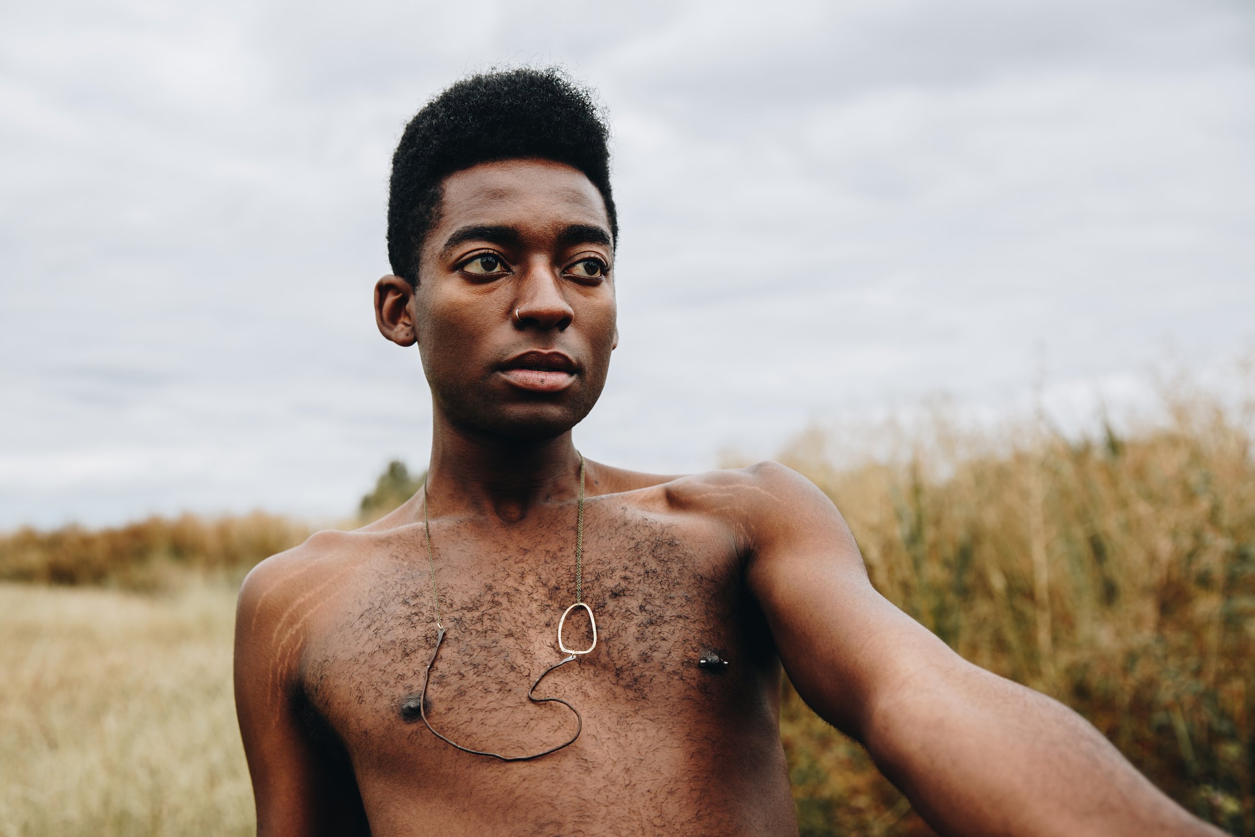 A shirtless young Black person with short  hair stands outdoors in a field of tall grass under a cloudy sky, wearing a unique necklace with an abstract wire design.