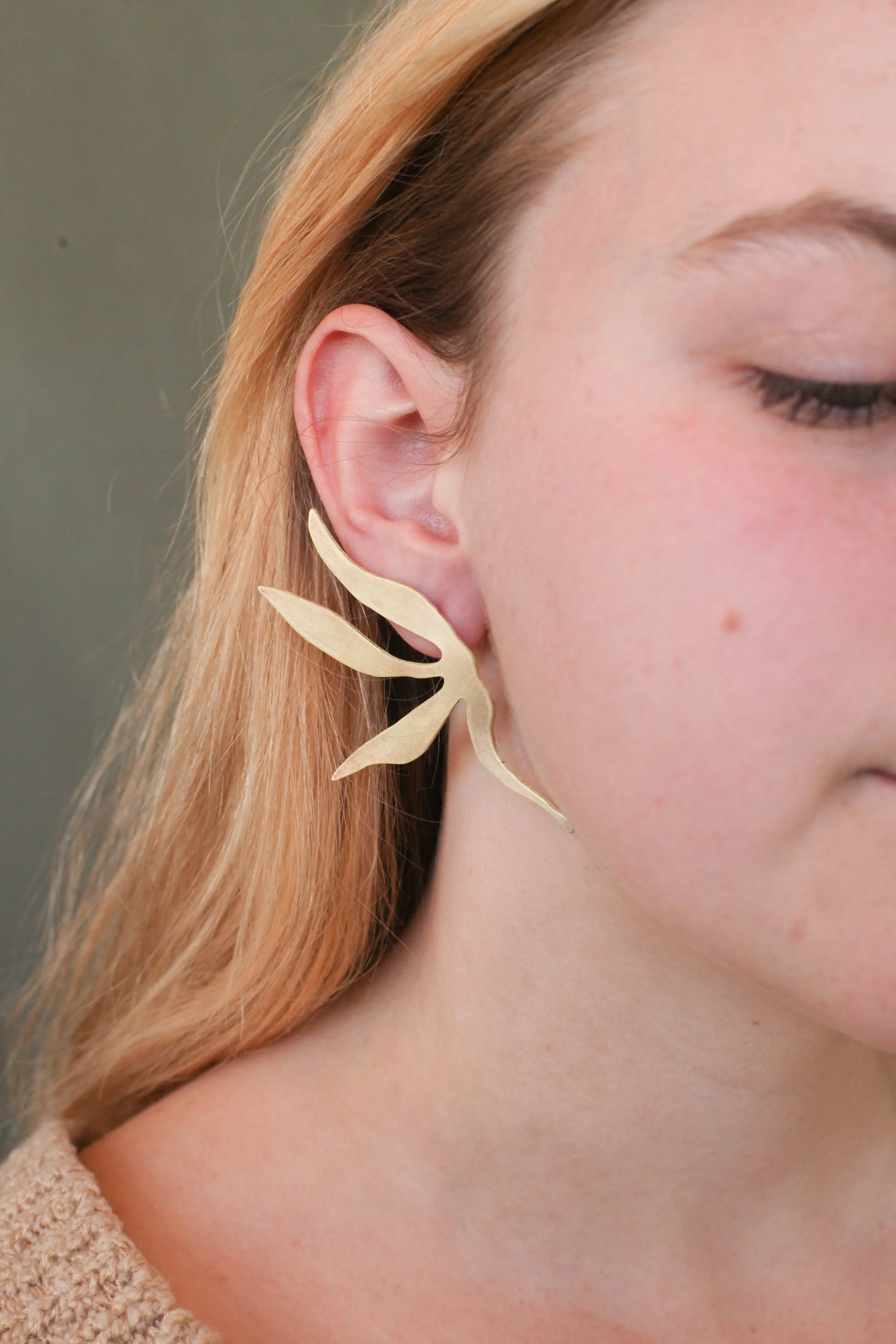 Close-up of a woman’s face showing her ear and part of her cheek, wearing a statement earring in the shape of a leaf.