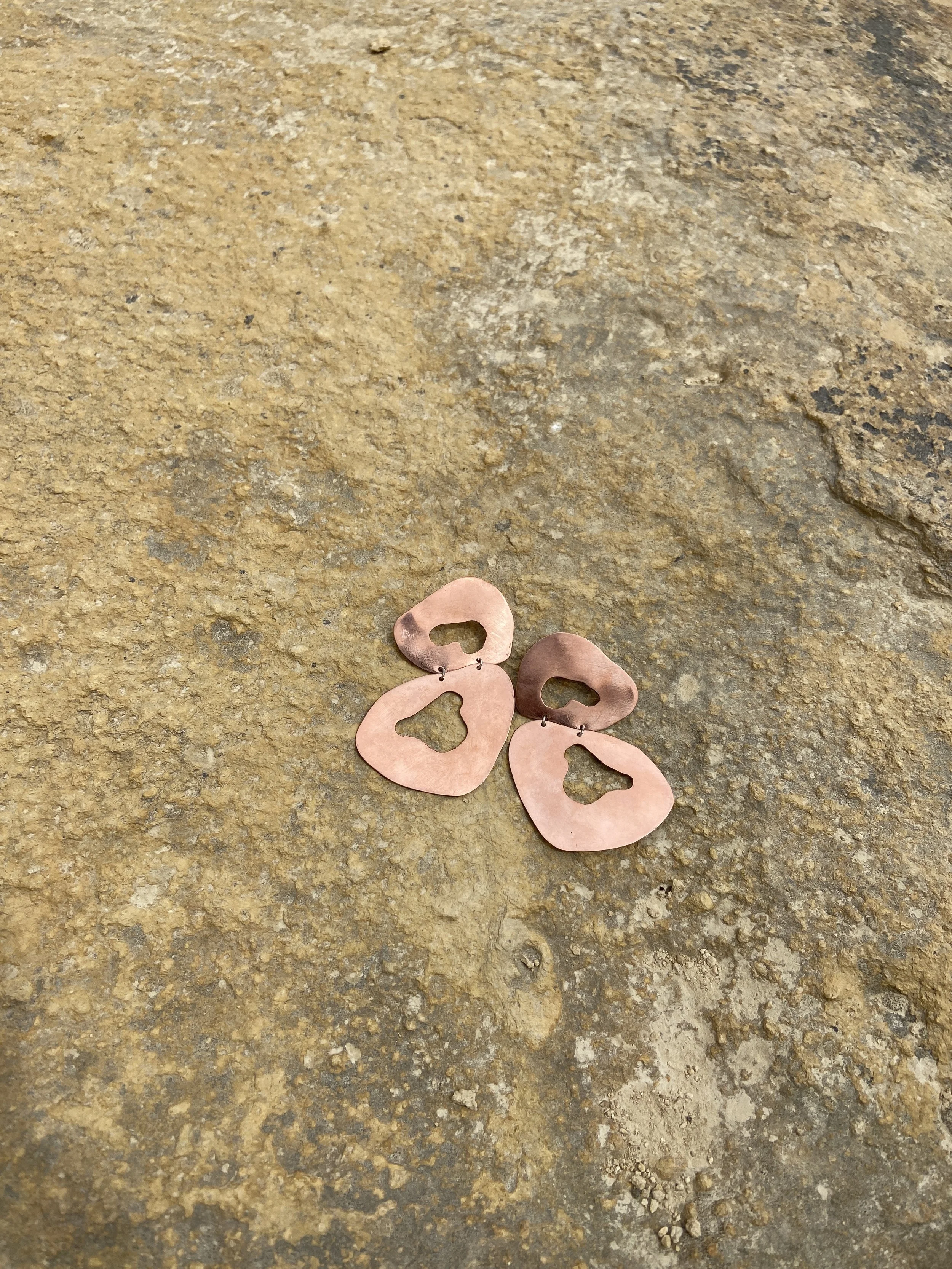 Pair of copper earrings with cutout shapes, resting on a rough, tan stone surface.
