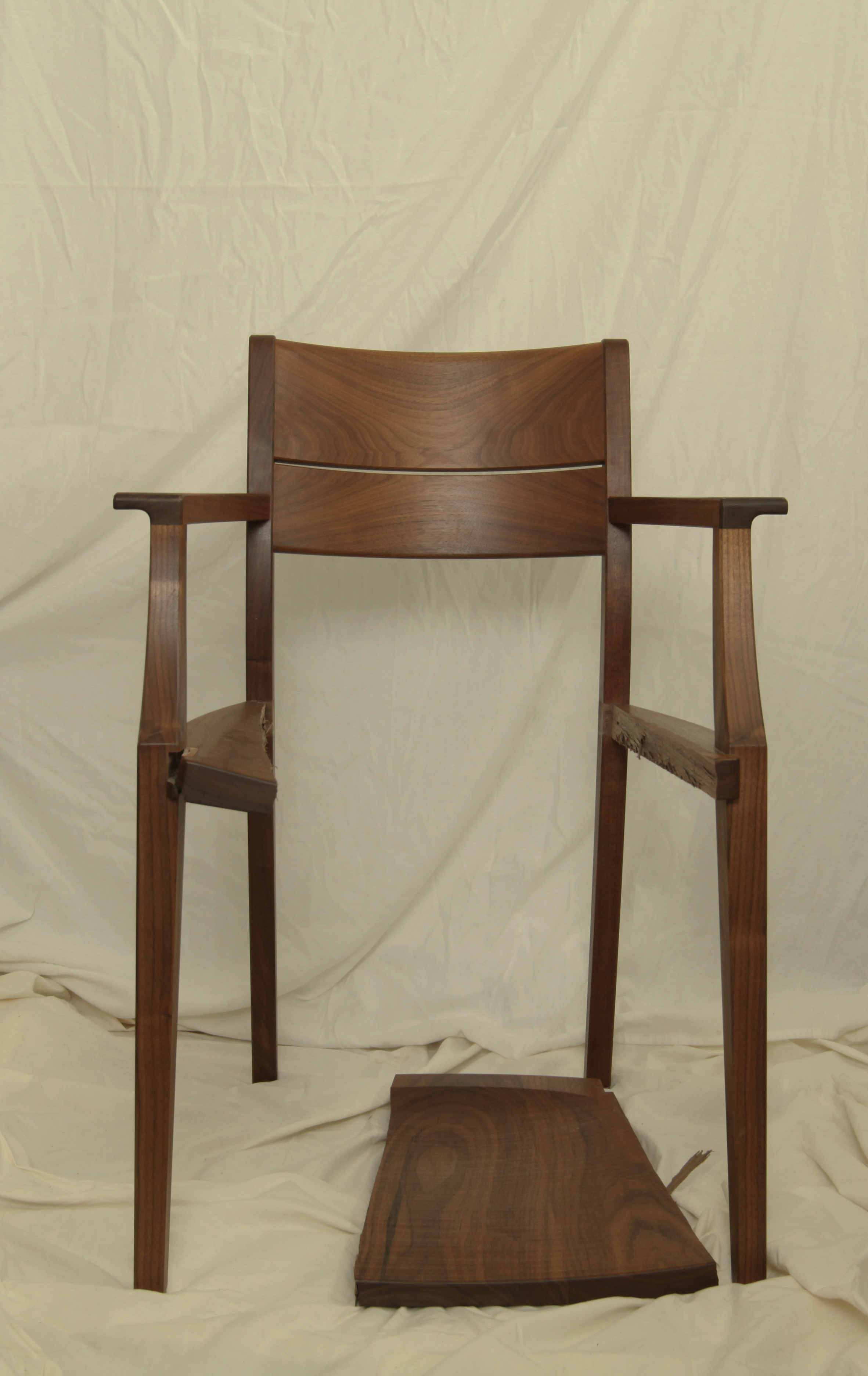 A broken wooden chair with no seat, showing the legs and arms, placed on a cream-colored fabric backdrop.