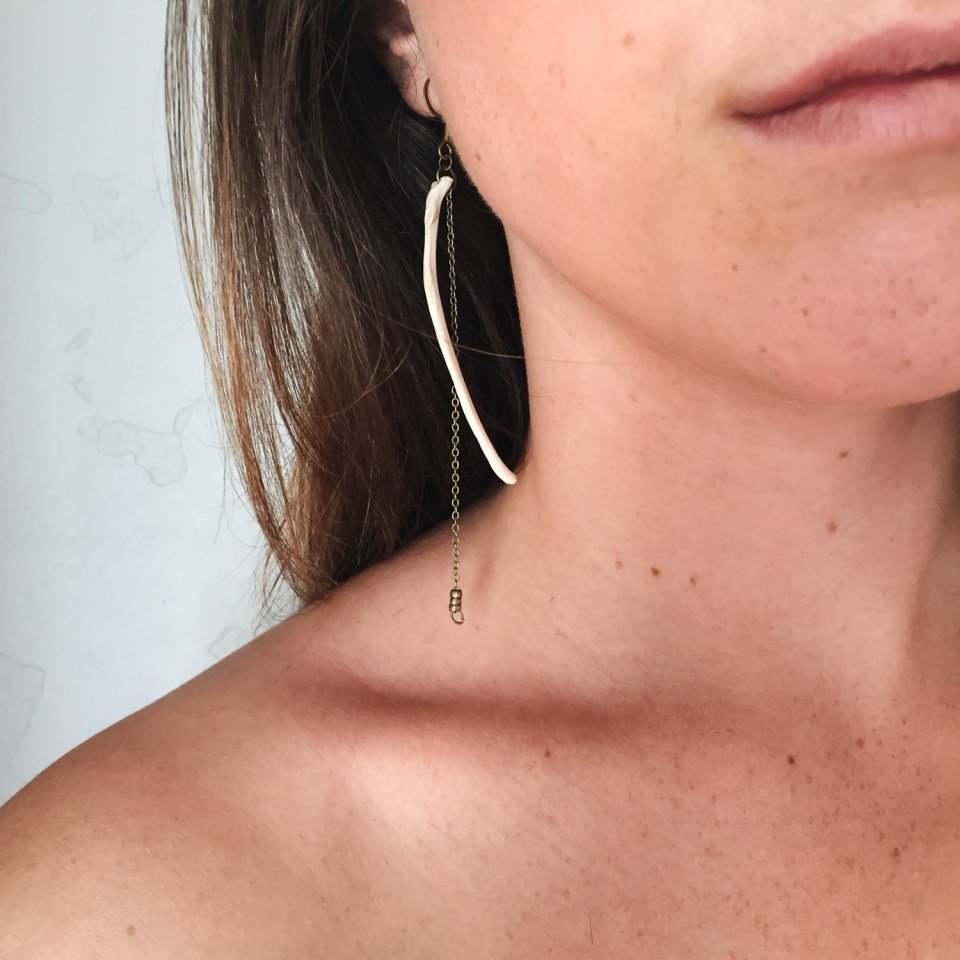 Close-up of a woman's left ear with a unique dangling earring, showing part of her cheek and shoulder. The earring features a long, curved white bone attached to a thin chain with a small spiral detail at the end.