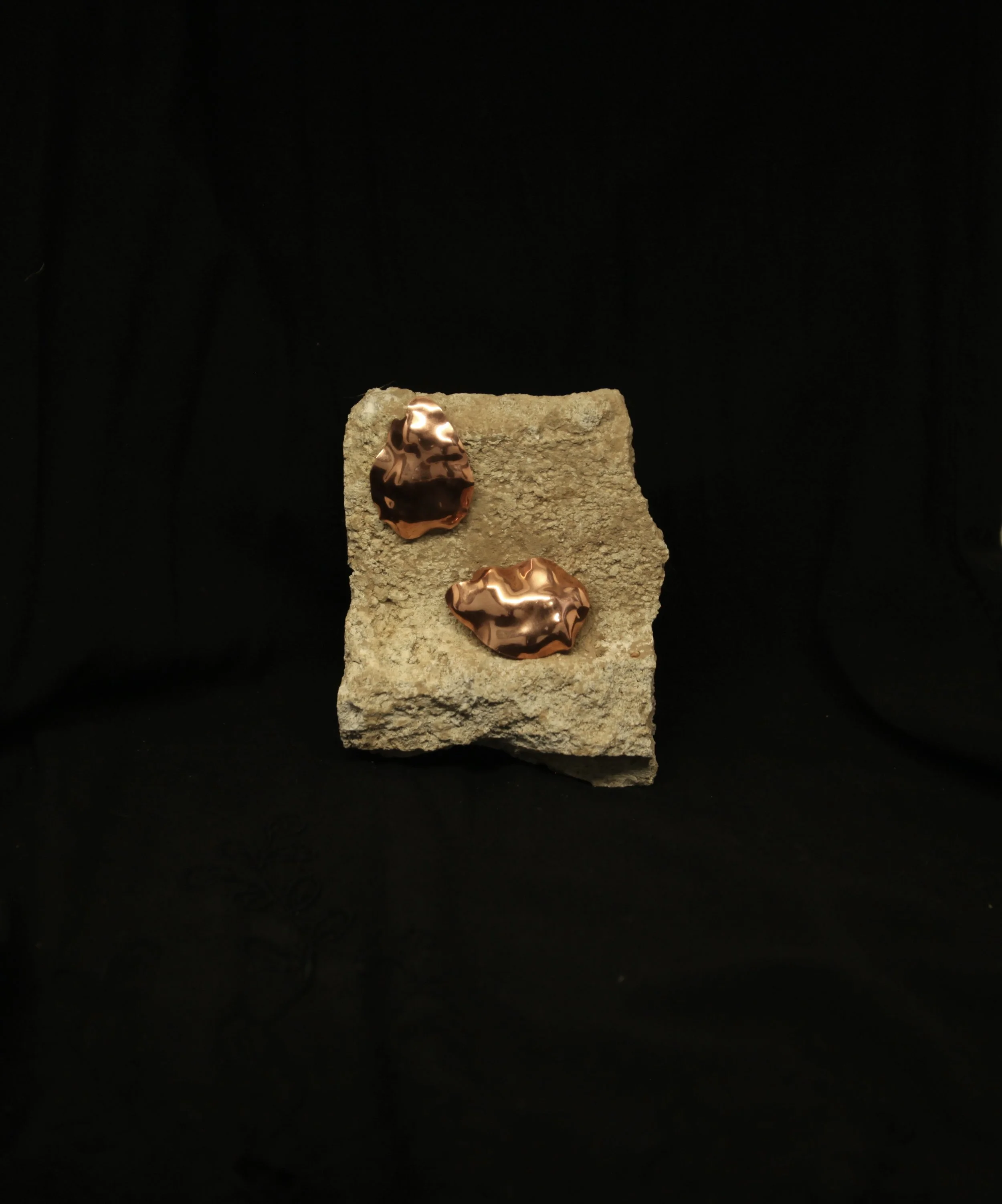 Two copper, unevenly shaped, bronze-colored objects on a rough stone slab against a black background.
