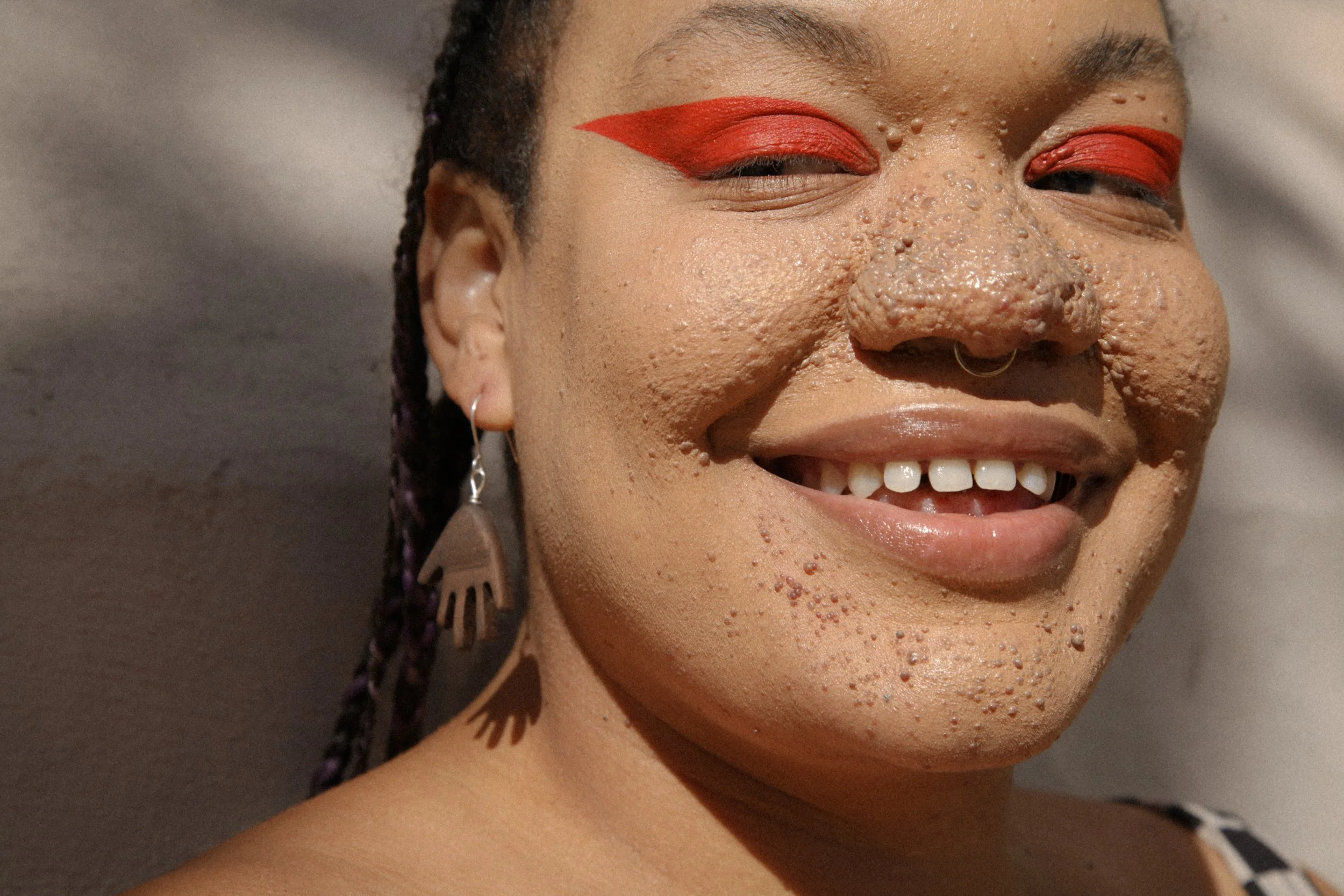 Close-up of a woman smiling with red eye makeup, wearing a unique earring and a septum piercing. She has textured skin and braided hair.