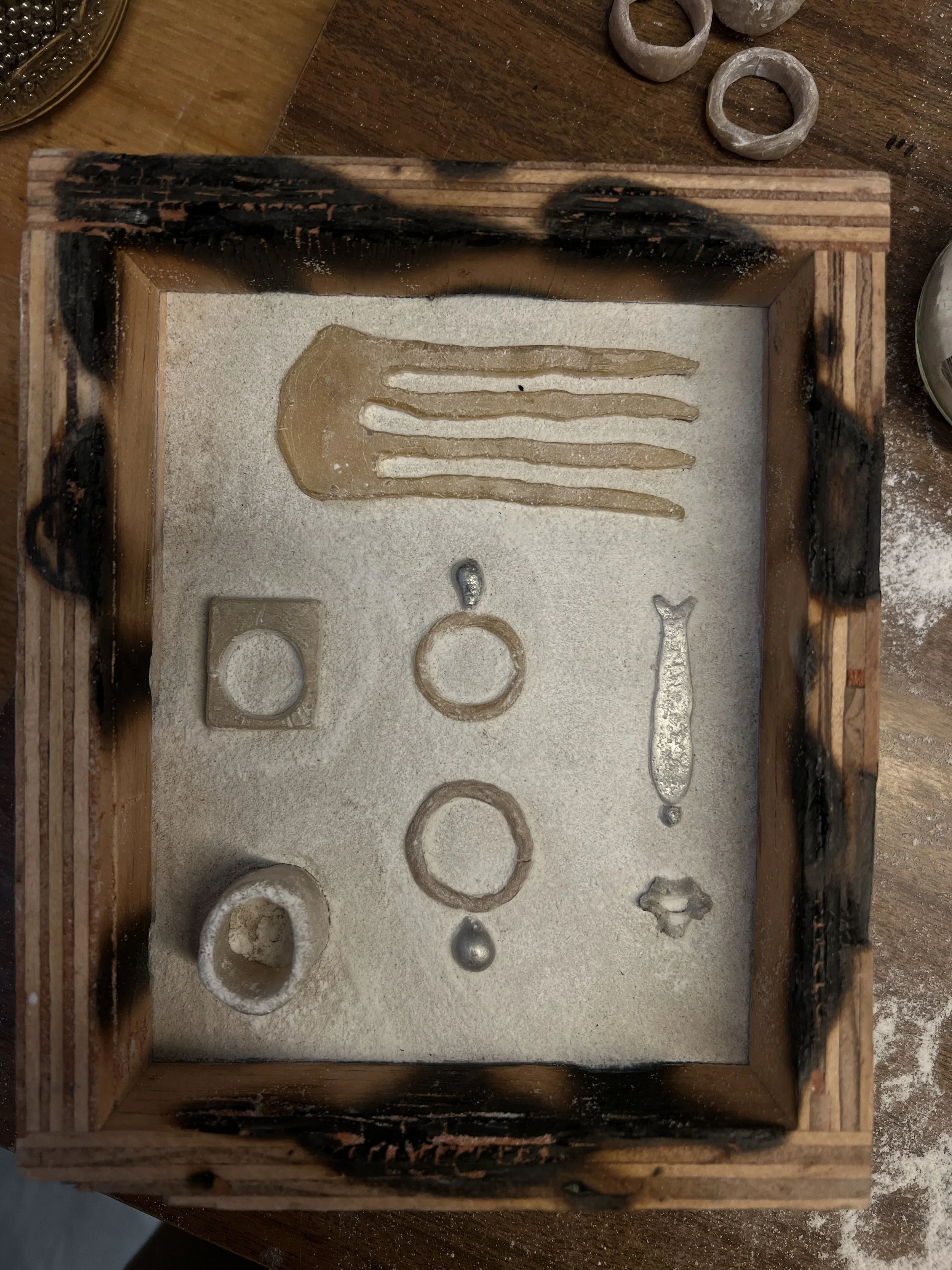 Various beeswax jewelry molds including rings, a pendant, and a small decorative item displayed on a bed of sand inside a wooden casting box
