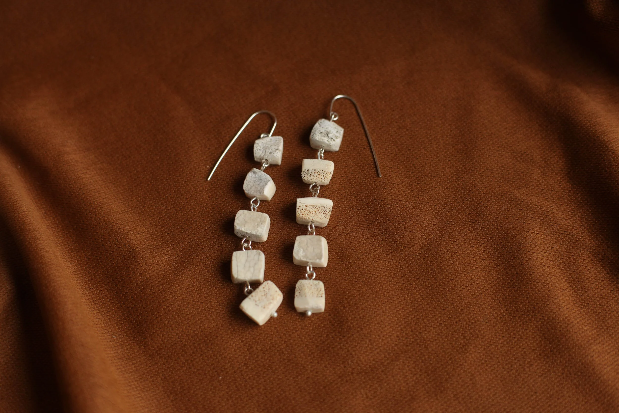 Pair of dangling earrings with square and rectangular white bone beads on a brown fabric background.