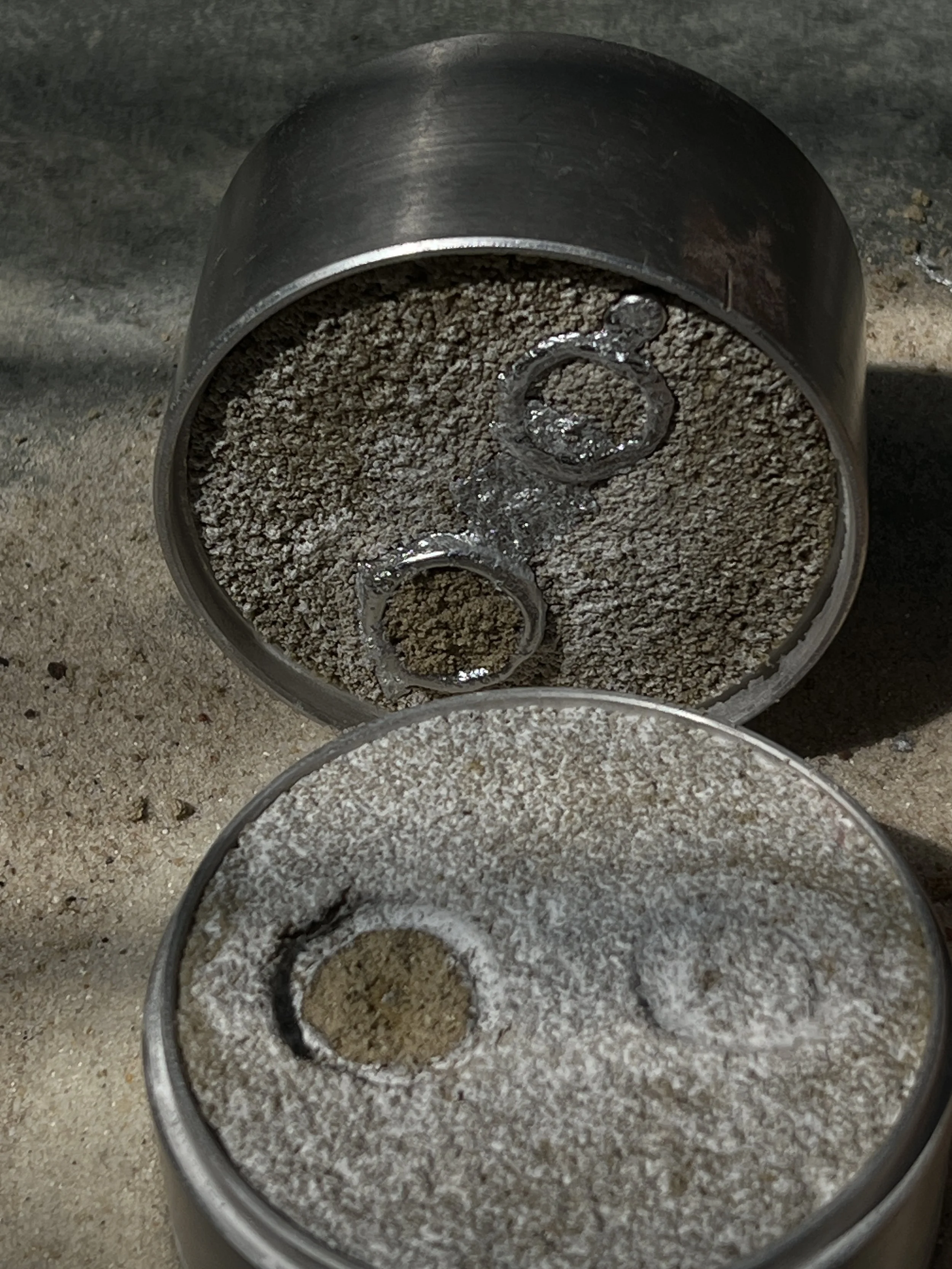 Two metal containers filled with a granular, sandy substance. The containers are open, and the granular material appears to be gritty and coarse. The background surface is rough and sandy as well.